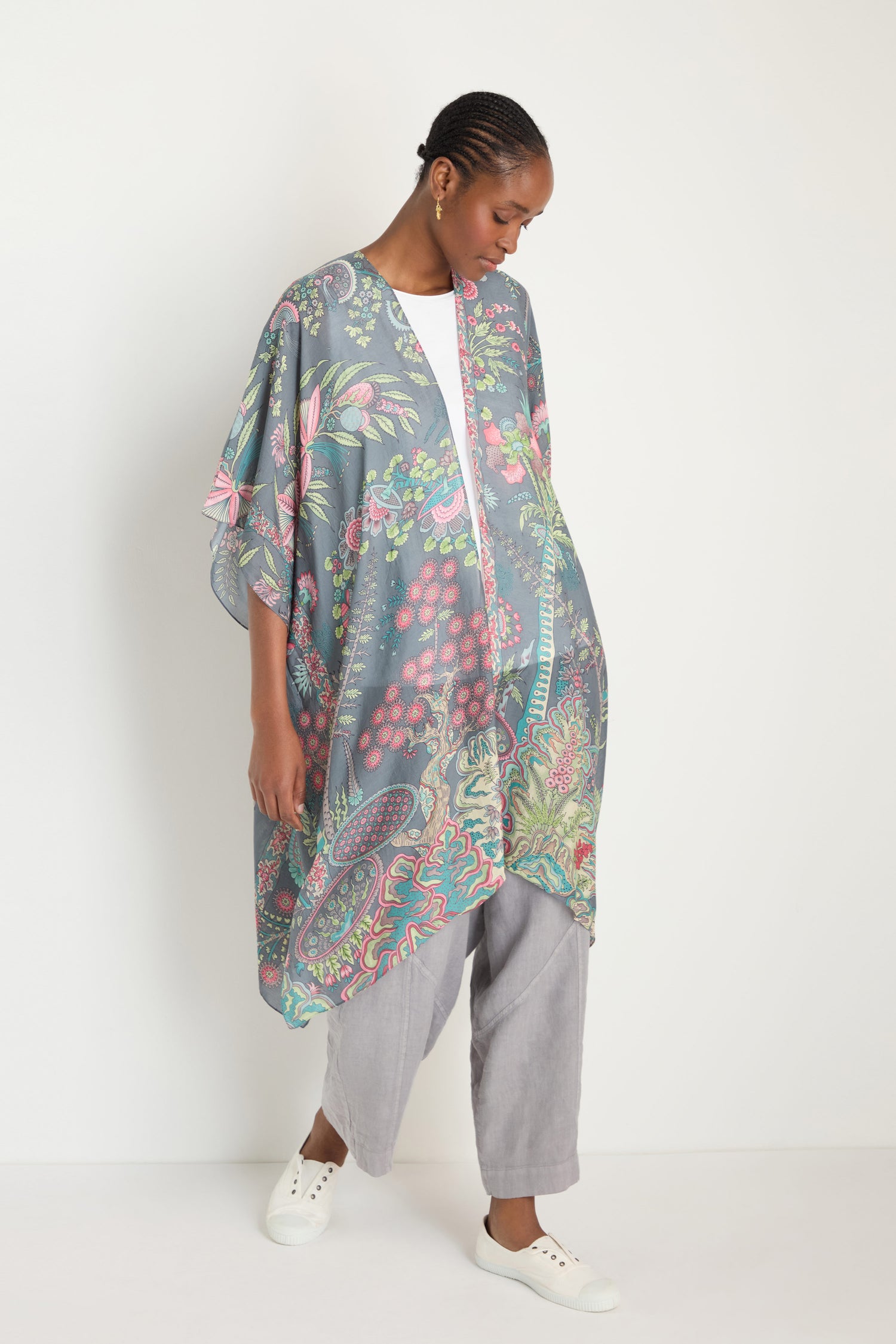A person stands against a white wall wearing the Boho Garden Print Throwover made from sustainable fabric, layered over a white shirt and gray pants, paired with white slip-on shoes.