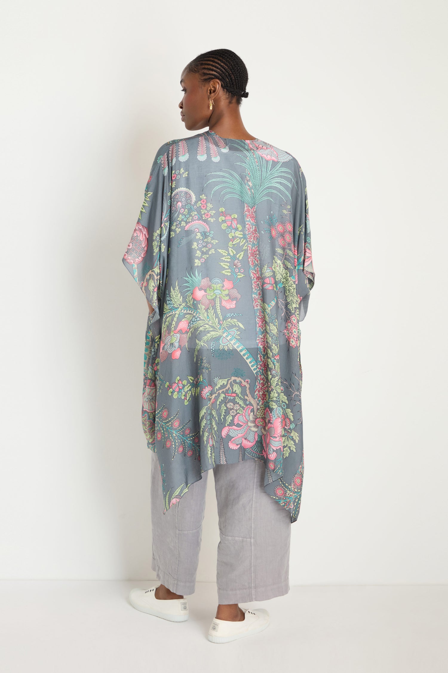 A person stands facing away, wearing the Boho Garden Print Throwover made from sustainable fabric over wide-legged gray pants and white slip-on shoes against a plain white background.