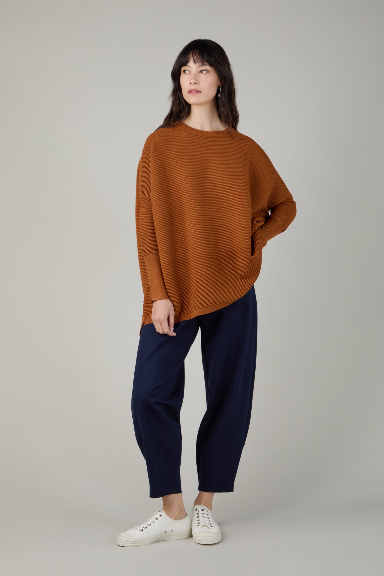 A woman stands against a plain background wearing a brown Ribbed Jumper, dark blue pants, and white sneakers, with one hand on her hip.