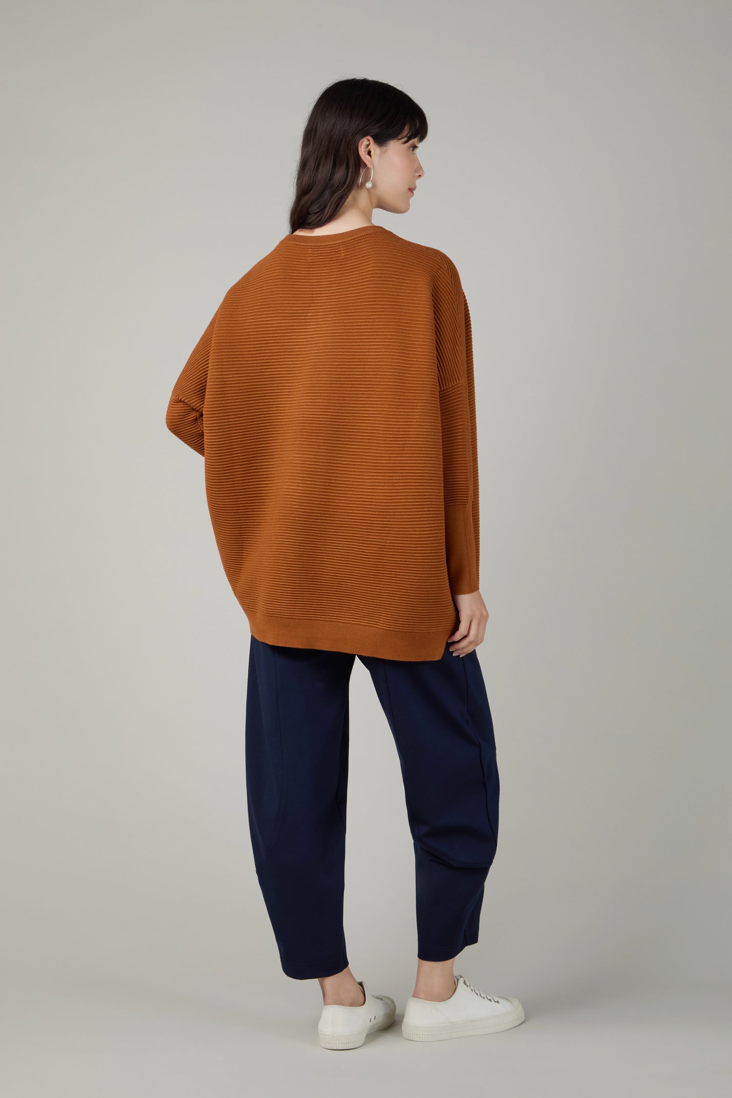 A person faces away wearing a loose Ribbed Jumper in brown, navy blue pants, and white sneakers against a plain gray background.