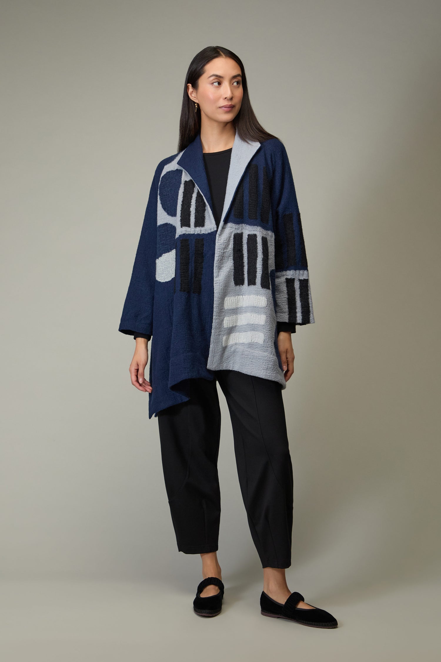 A woman stands wearing the Spot and Dash Wool Jacket in navy and gray abstract pattern from Raga, paired with black pants and black flat shoes against a plain background.