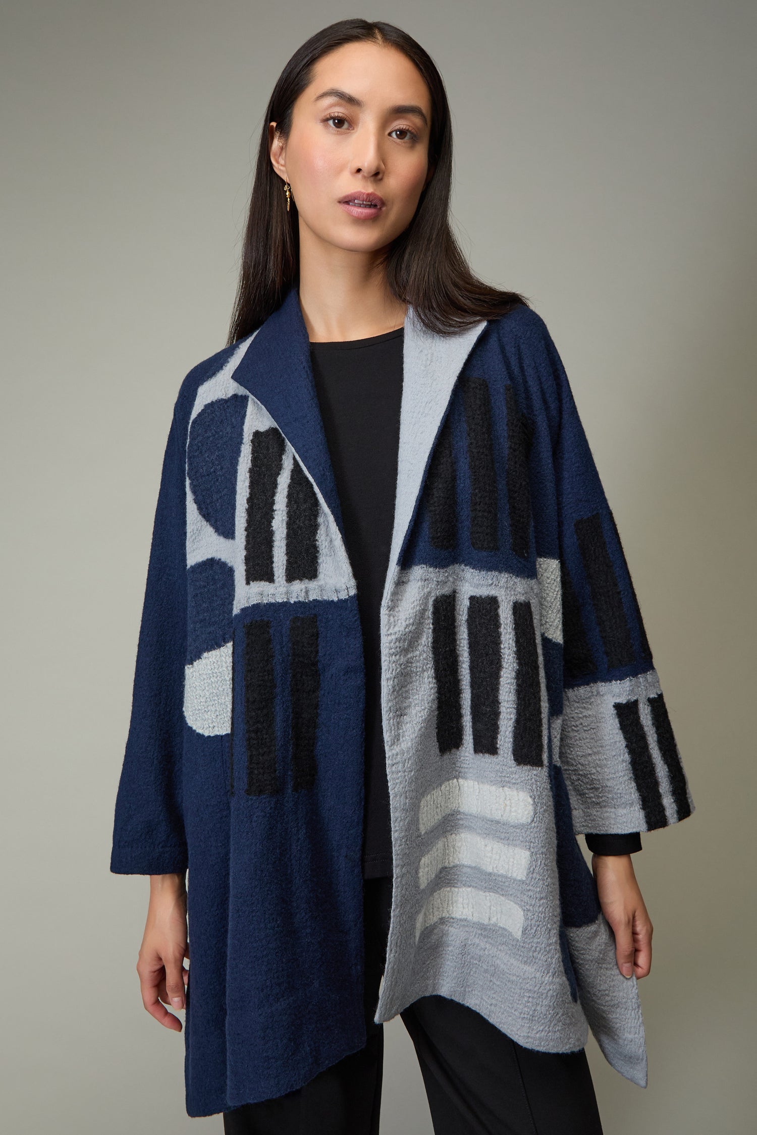 A woman stands against a plain background wearing a Spot and Dash Wool Jacket in abstract patterns of dark blue, light blue, and black over a black top and pants.