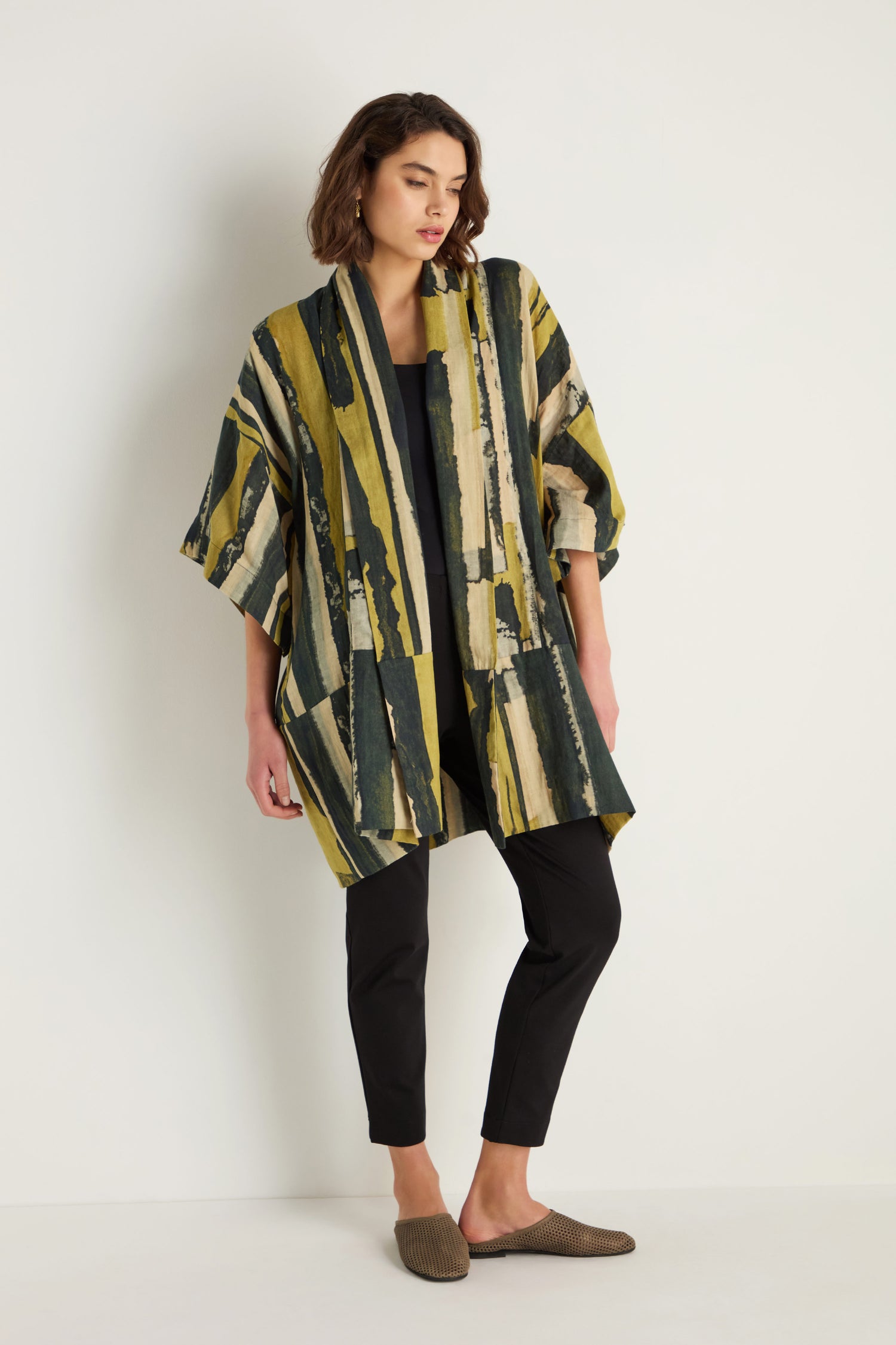 A woman stands against a white wall wearing the Oversized Cotton Kimono with a painterly print, paired with a black top, black pants, and brown slip-on shoes.