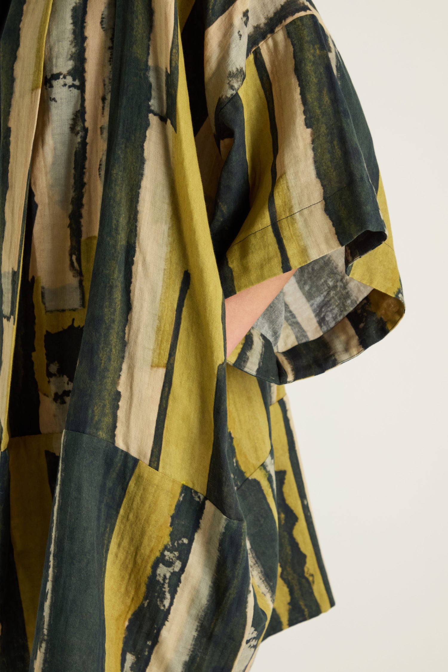A close-up of the Oversized Cotton Kimono with wide sleeves displays a painterly print in green, yellow, beige, and dark blue. A hand is partially visible in the pocket.