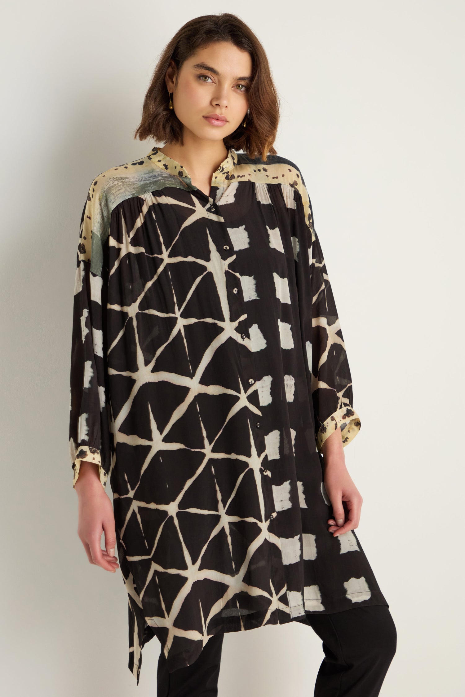 A woman wears the Shibori Print Long Shirt—a relaxed fit, long black shirt with beige geometric patterns in a cupro-viscose blend—standing against a plain light background.