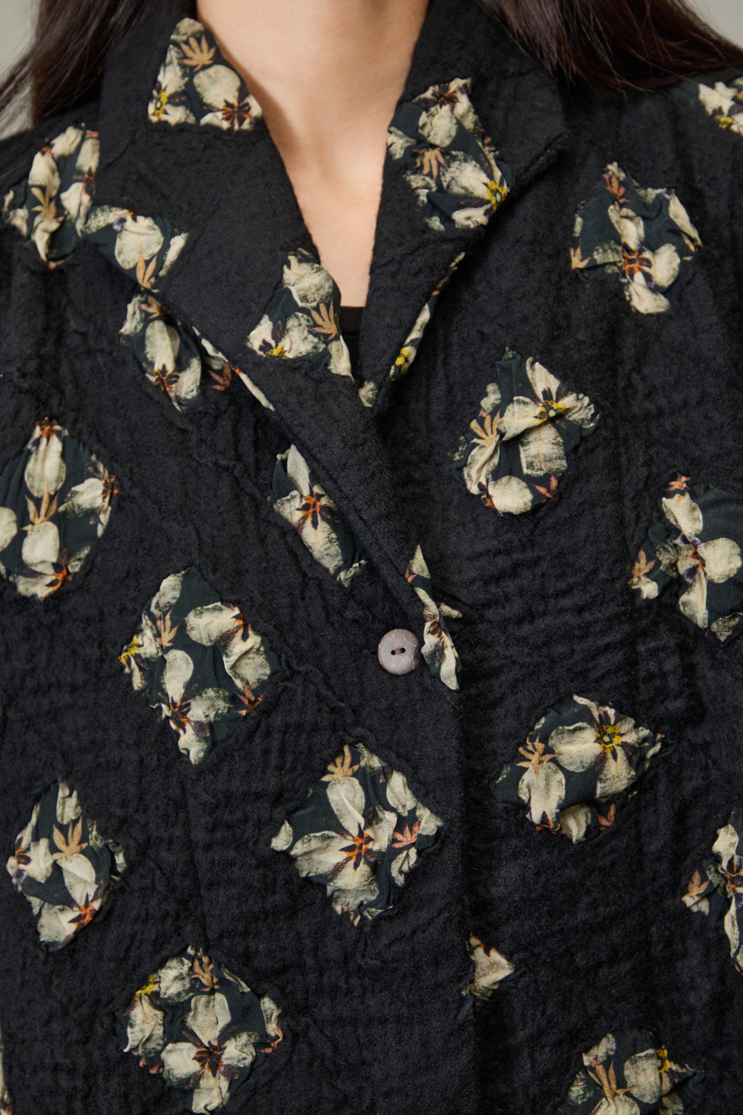 Close-up of a person wearing the Textured Flower Wool Jacket—a black jacket with cream flowers and green leaves in square clusters, giving a subtle quilted effect.