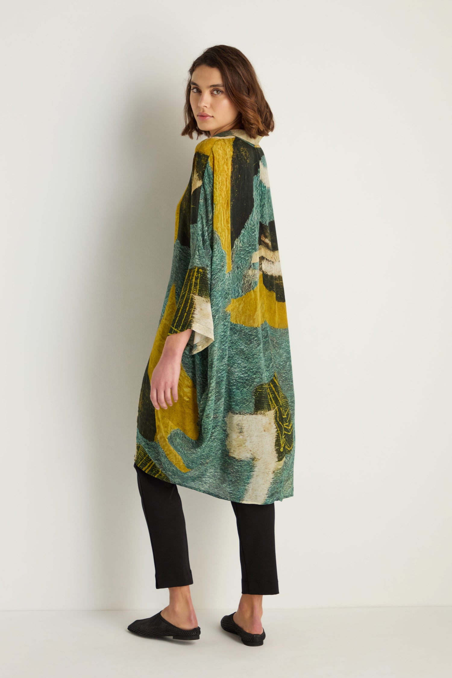 A person stands sideways against a plain white background, wearing the Ochre Print Shirt Dress with black pants and black shoes.