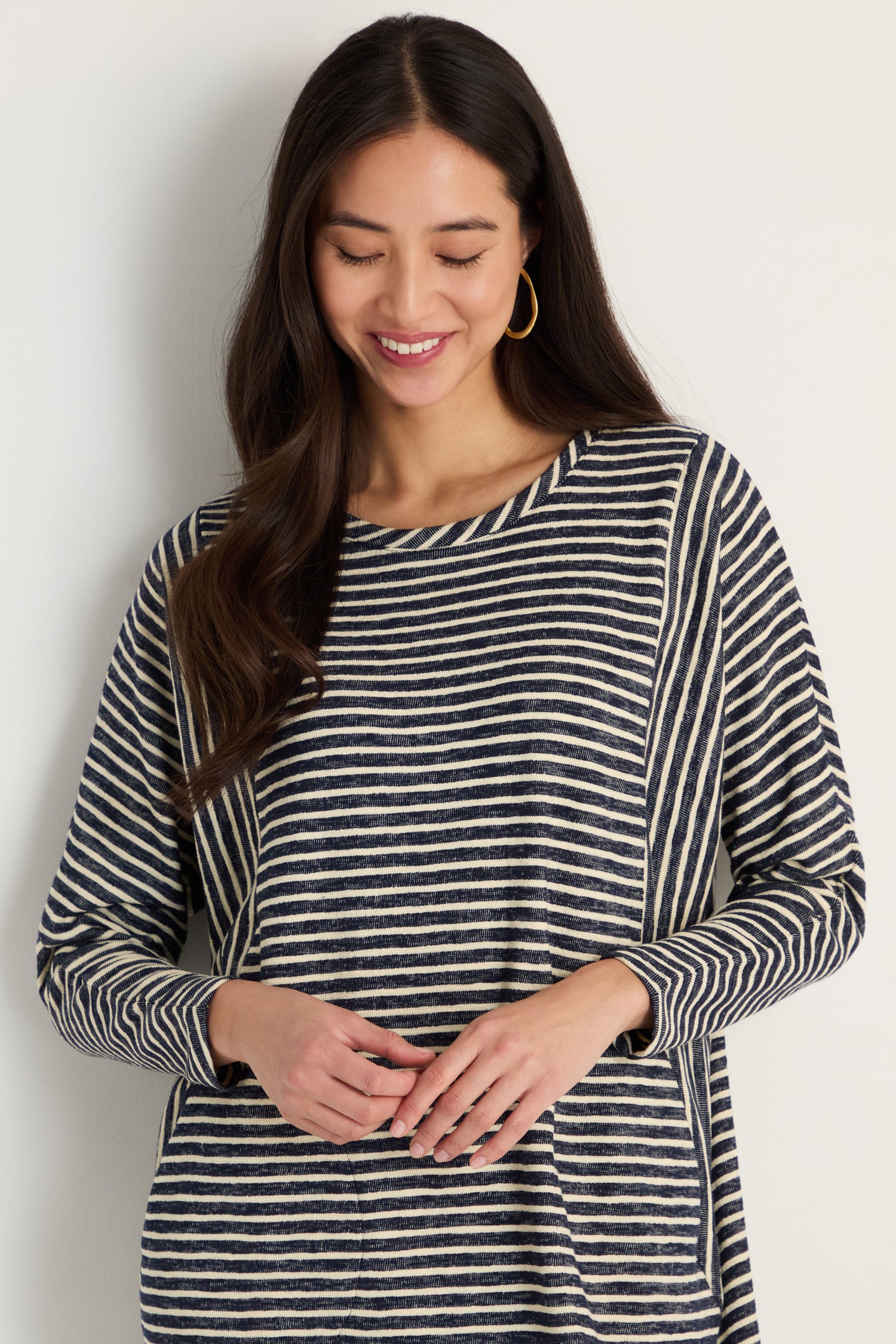 A woman with long dark hair and hoop earrings smiles as she looks down, modeling the Textured Stripe Jersey Dress against a light background.