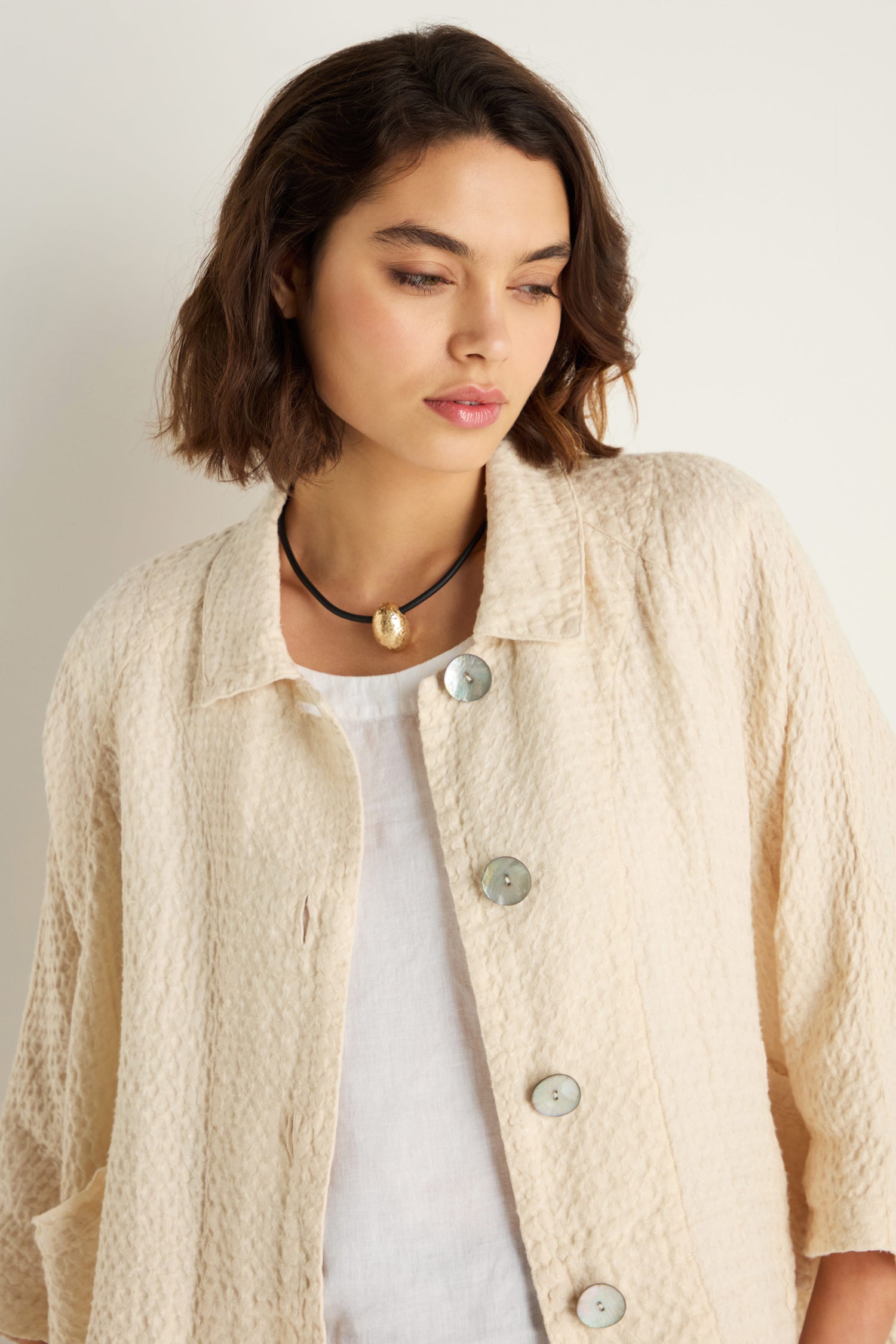 A woman with short brown hair stands against a plain background, wearing a textured cream button-up jacket over a white top and the Hammered Pebble Necklace, which features a gold pendant.