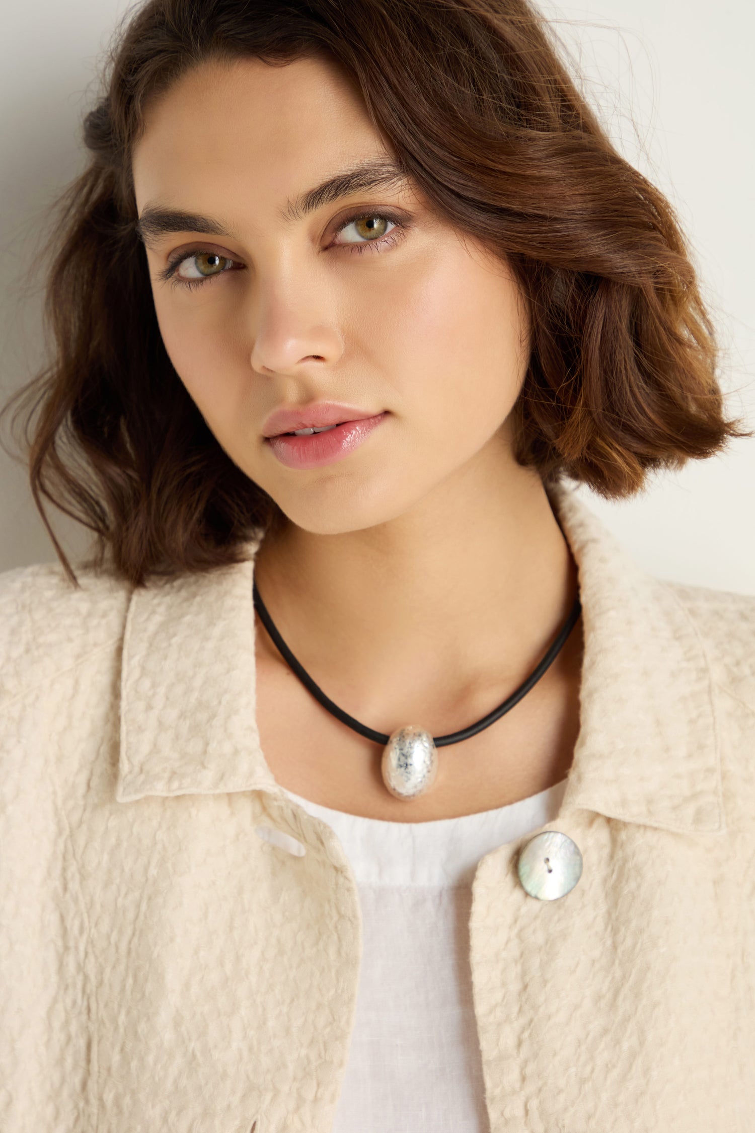 Woman with short brown hair wears a textured cream jacket, white top, and the Hammered Pebble Necklace—a black rubber cord choker with a silver pendant—standing against a light background.