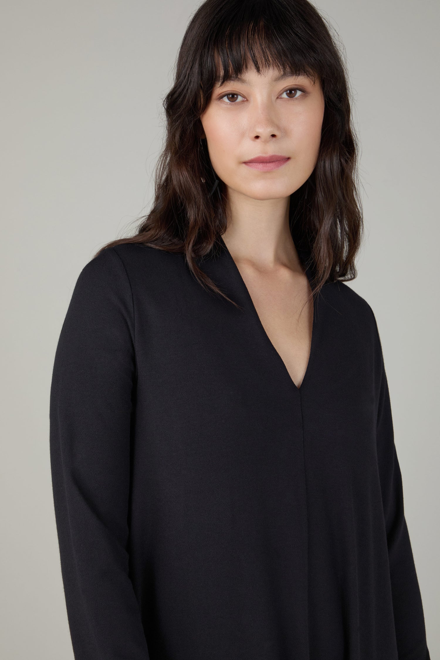 A woman with long dark hair and bangs stands against a plain light gray background, wearing the Fluid Crepe V Neck Black Bubble Dress.
