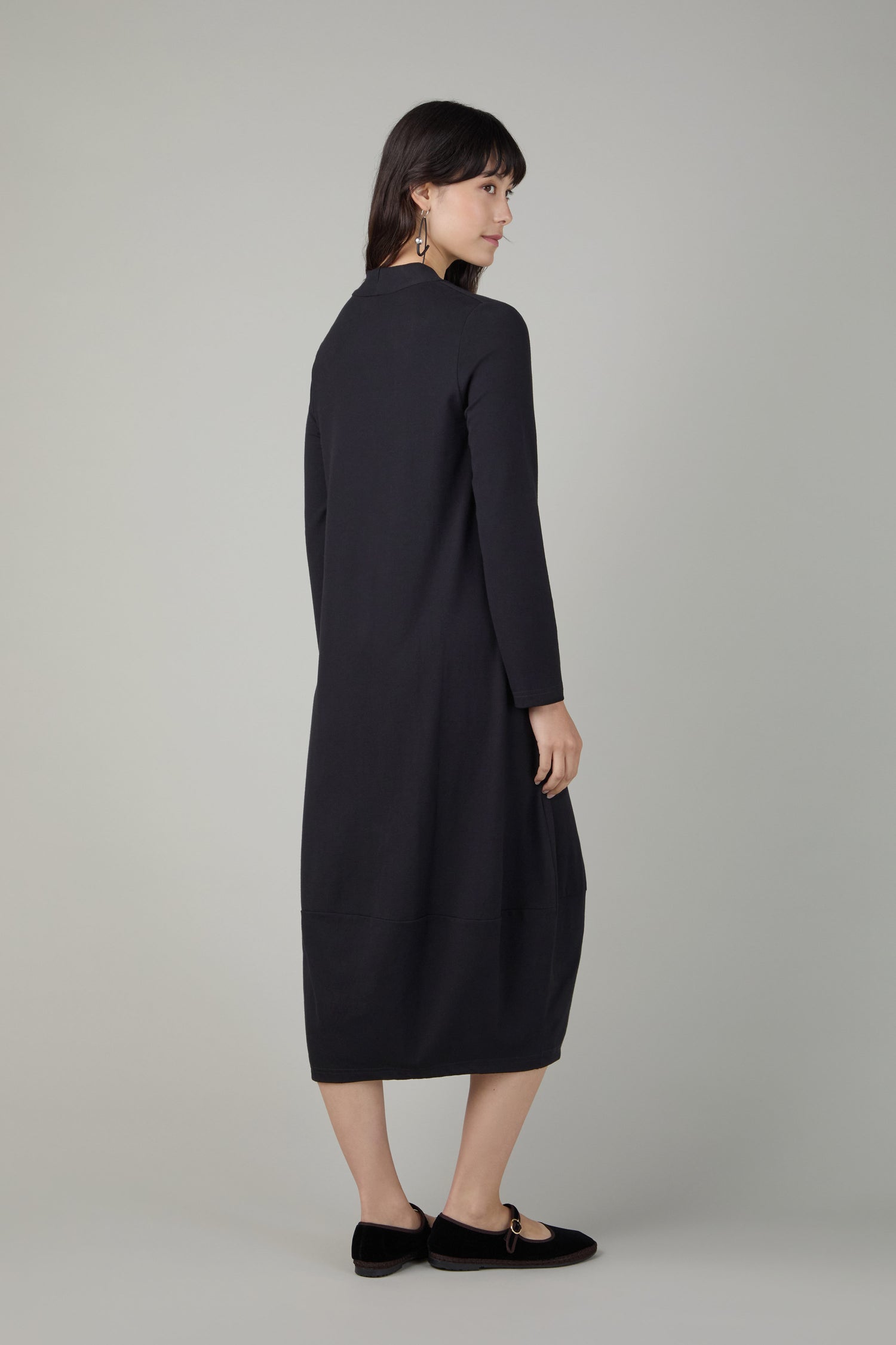 A woman faces slightly away, wearing the Fluid Crepe V Neck Black Bubble Dress with black flats, set against a plain light gray background.