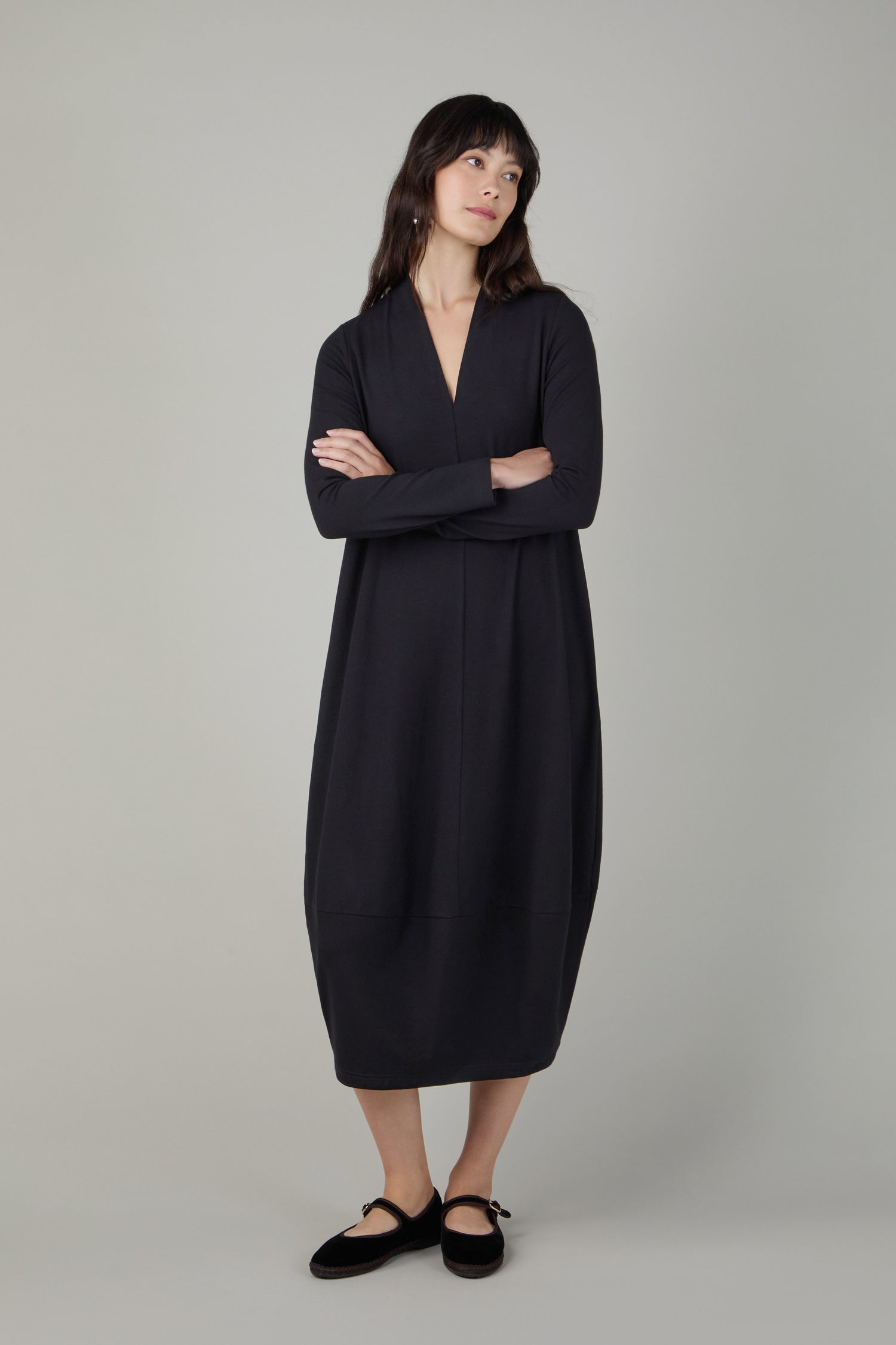 A woman stands with arms crossed, wearing the Fluid Crepe V Neck Black Bubble Dress—a loose, long-sleeve black crepe jersey piece with a subtle bubble silhouette—paired with black flats against a plain light gray background.