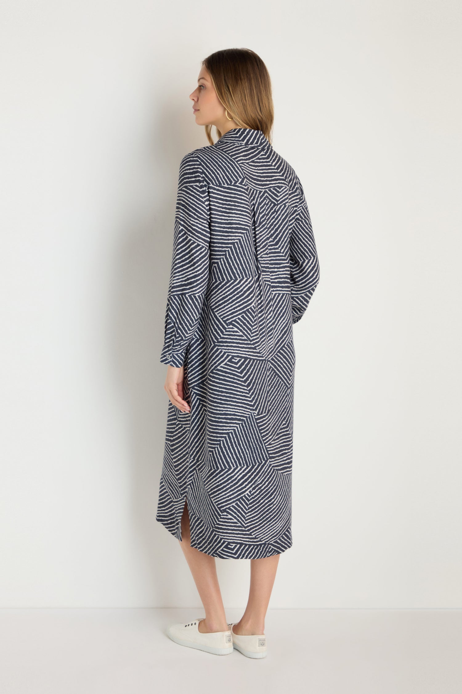 A woman faces away, wearing the Linear Prism Shirt Dress with long sleeves and a geometric print, paired with white slip-on shoes against a plain white background.