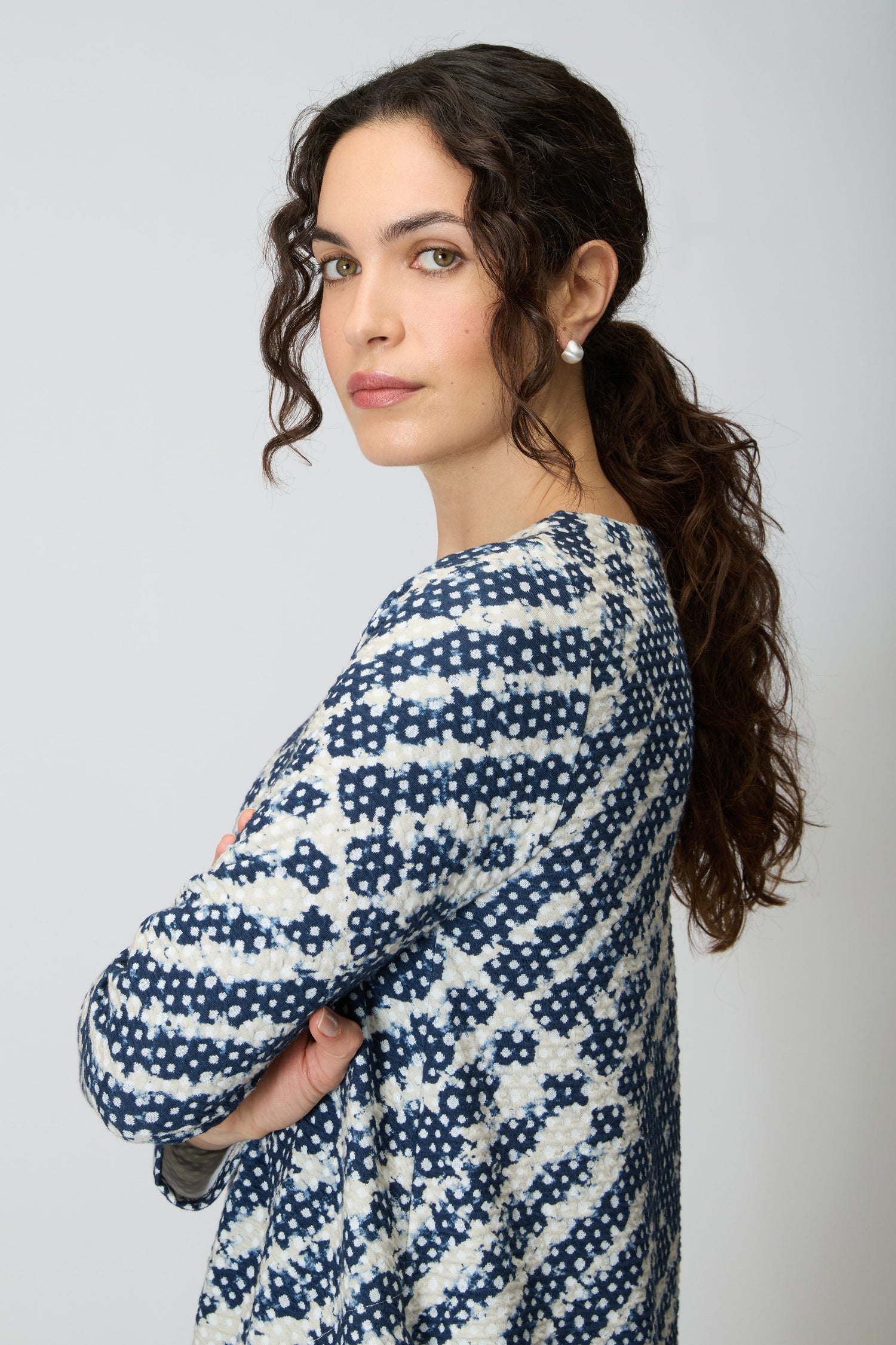 A woman with long curly brown hair in a low ponytail wears the Diamond Tie Dye Jersey Bubble Dress, standing with arms crossed and looking at the camera against a plain background.