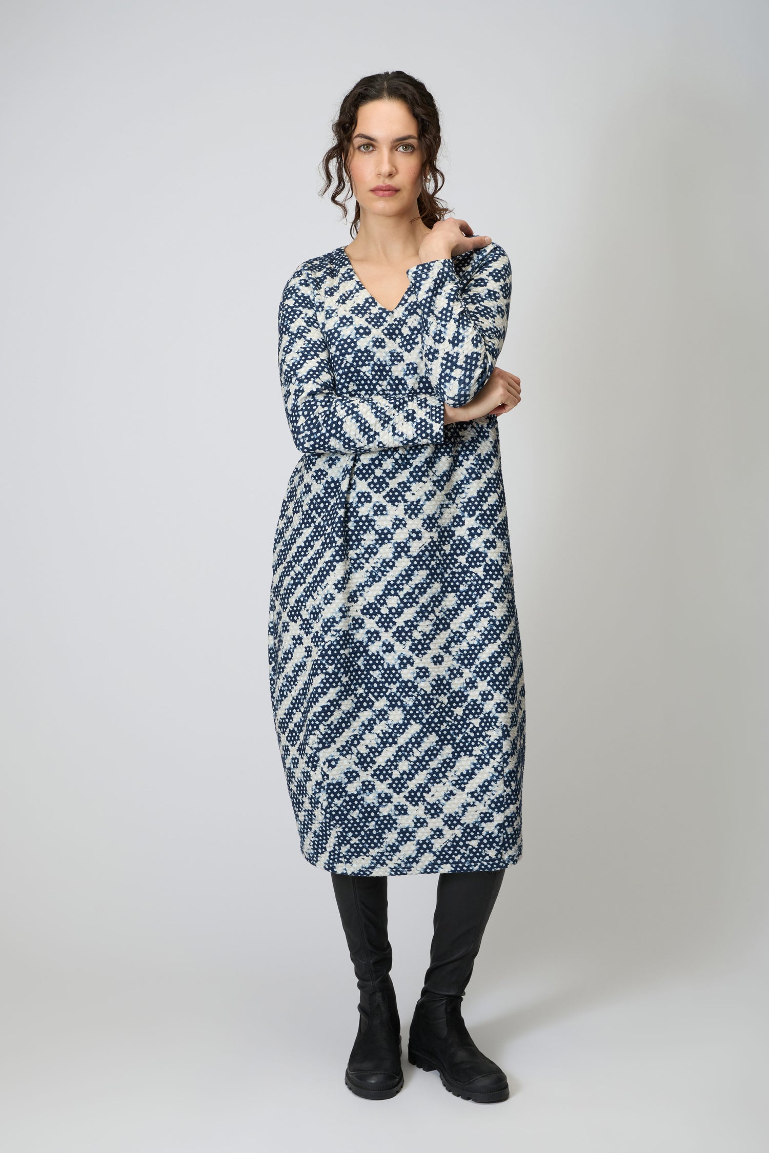 A woman with curly hair and a neutral expression stands against a plain background wearing the Diamond Tie Dye Jersey Bubble Dress, styled with a belt, black leggings, and boots.