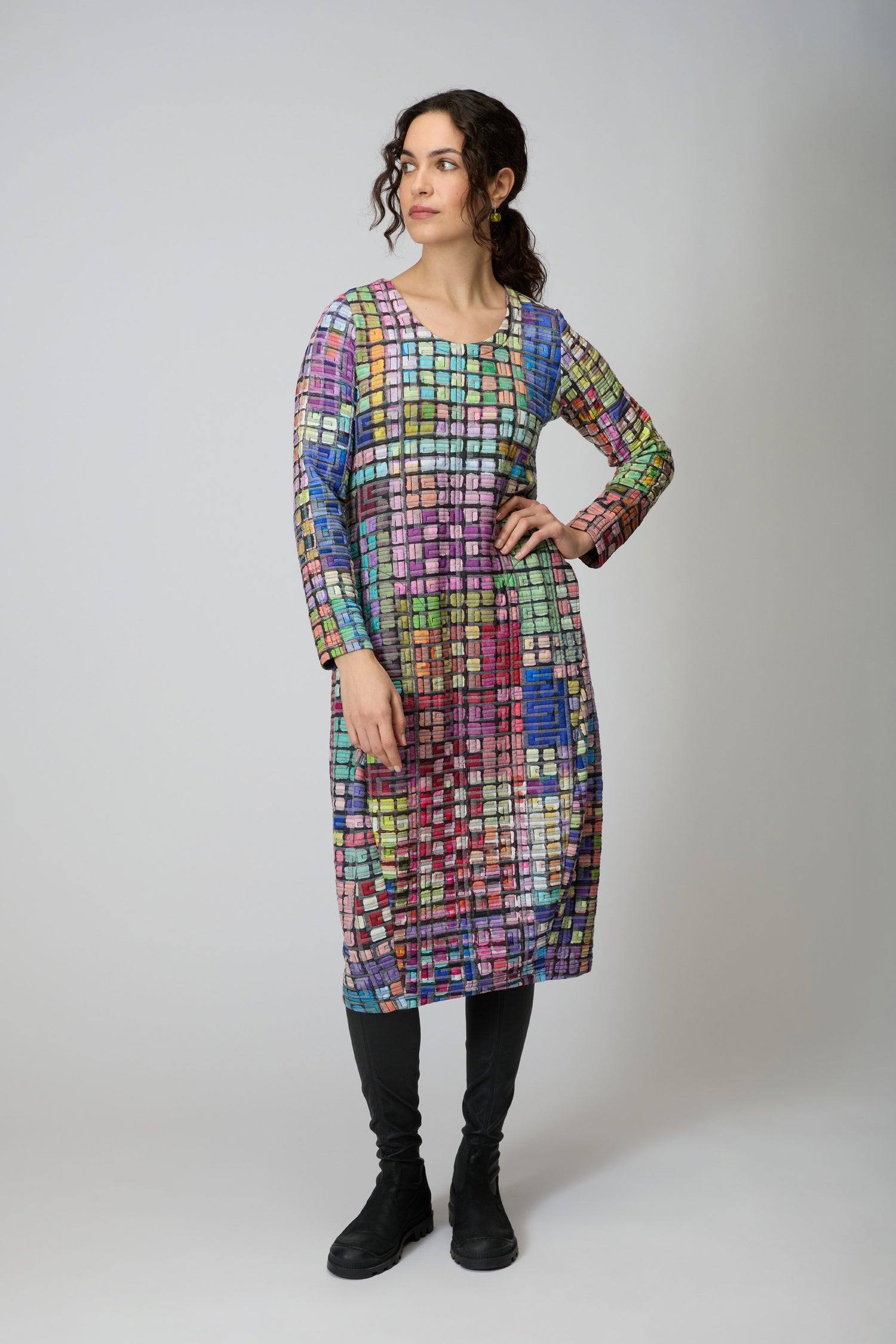 A woman faces slightly left, wearing the Coloured Glass Tiles Jersey Bubble Dress with a vibrant mosaic pattern, paired with black leggings and shoes, set against a neutral background.