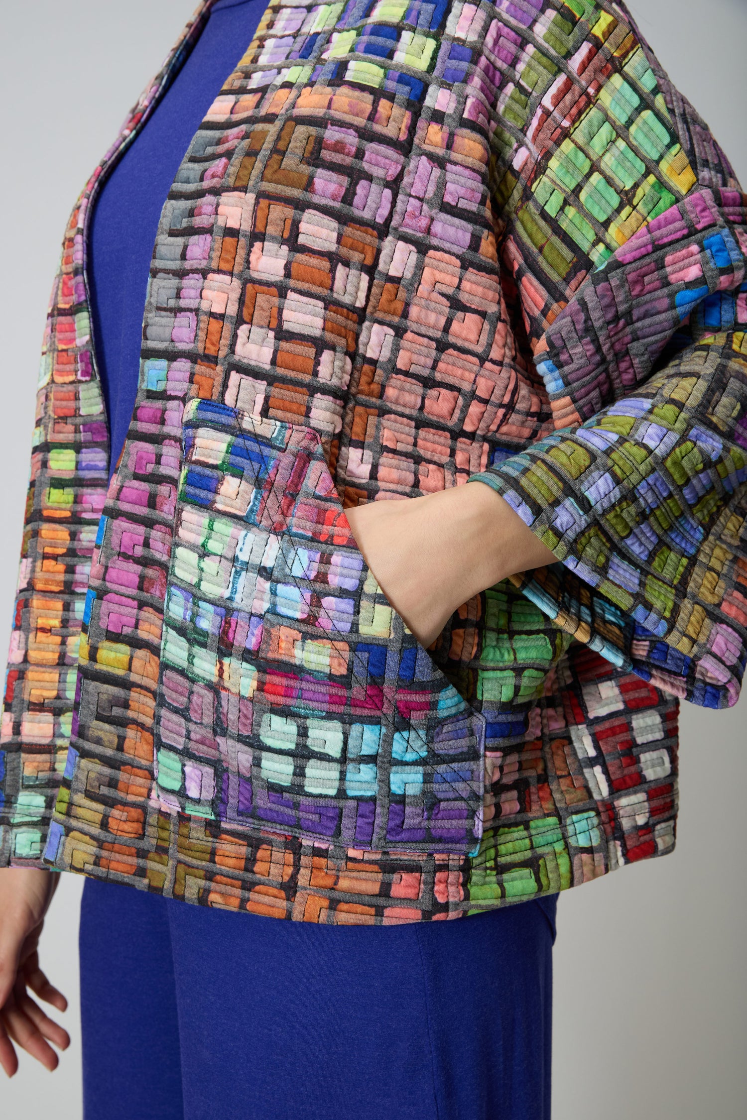 A person wears the Coloured Glass Tiles Jersey Jacket, featuring a multicolored grid design like colored glass tiles, over a blue outfit; their hand is tucked into one of the jacket’s front pockets.