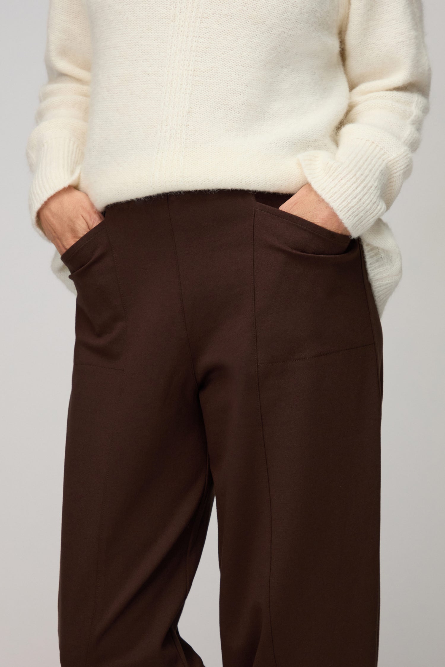 A person stands with hands in their pockets, wearing a white knit sweater and Chocolate Ponte Bubble Trousers featuring a sculptural bubble shape.