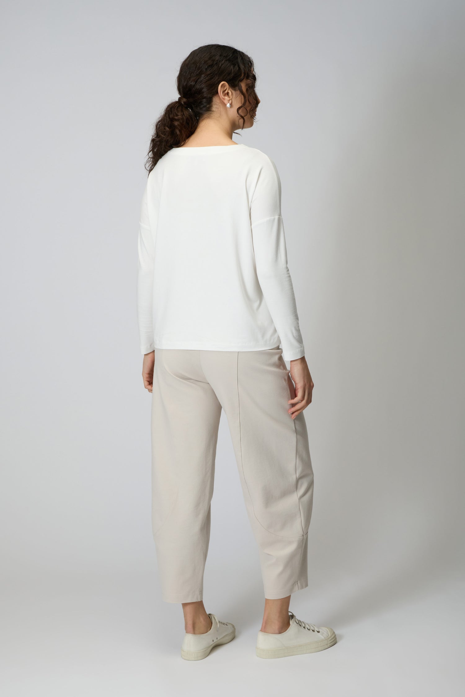 A person stands facing away, wearing a white long-sleeve top, Ecru Ponte Bubble Trouser in a bubble leg silhouette, and white sneakers against a plain light background. Made in England.