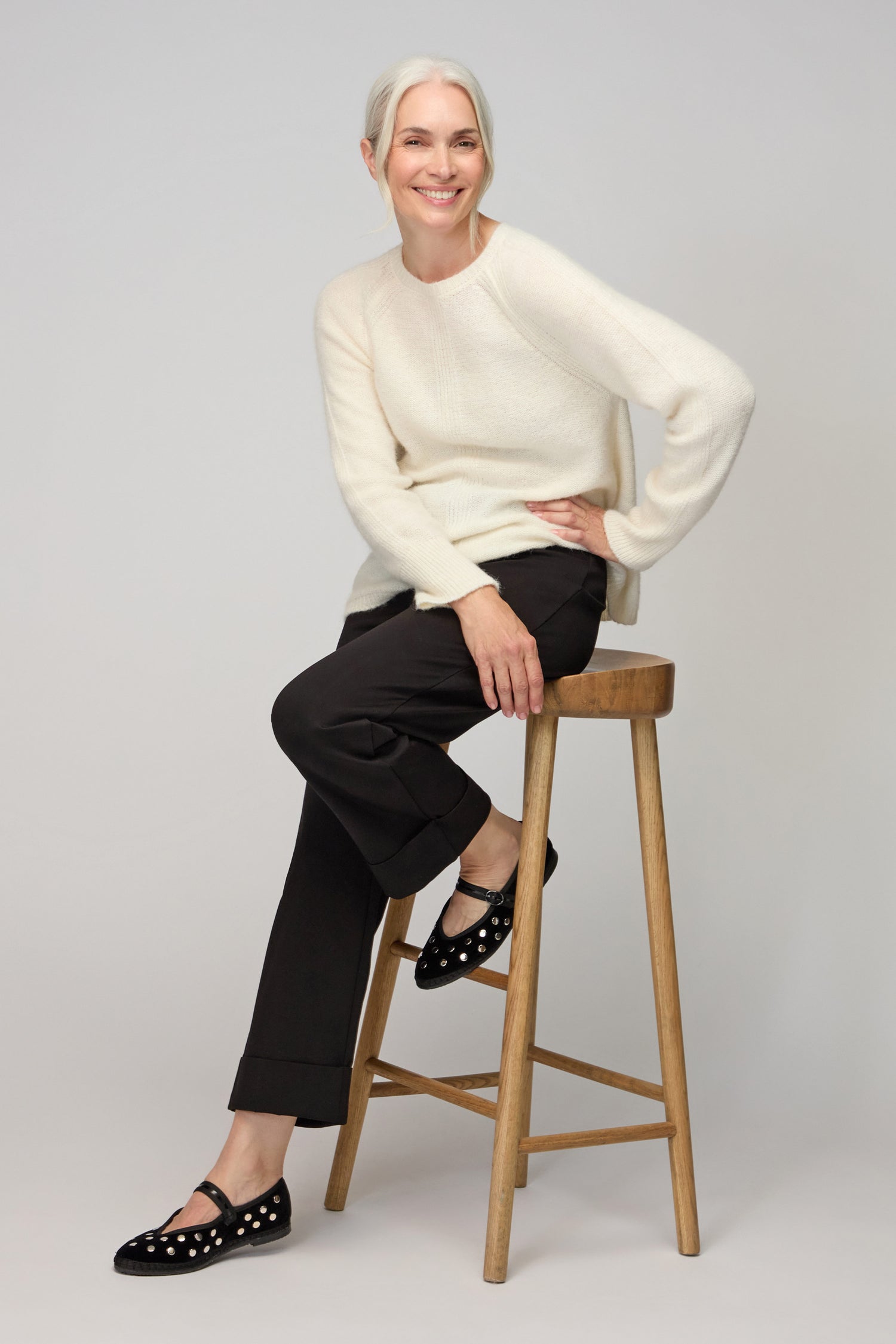 A woman with gray hair in a white sweater and Black Ponte Straight Trousers sits on a wooden stool, smiling, against a plain light background.