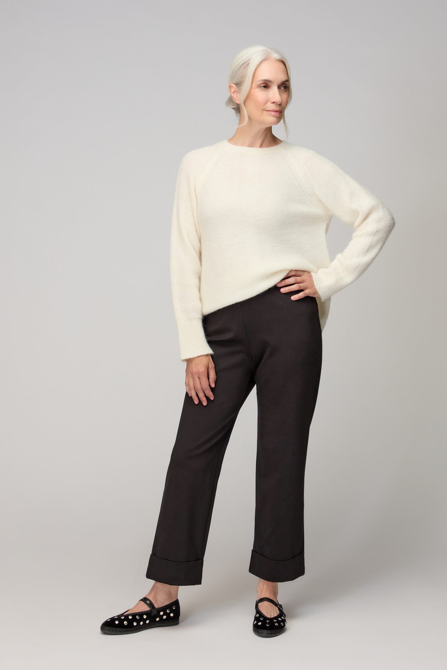A woman with light hair in a low ponytail wears a white sweater and the Black Ponte Straight Trousers, paired with black loafers featuring decorative details, as she stands against a plain light background.