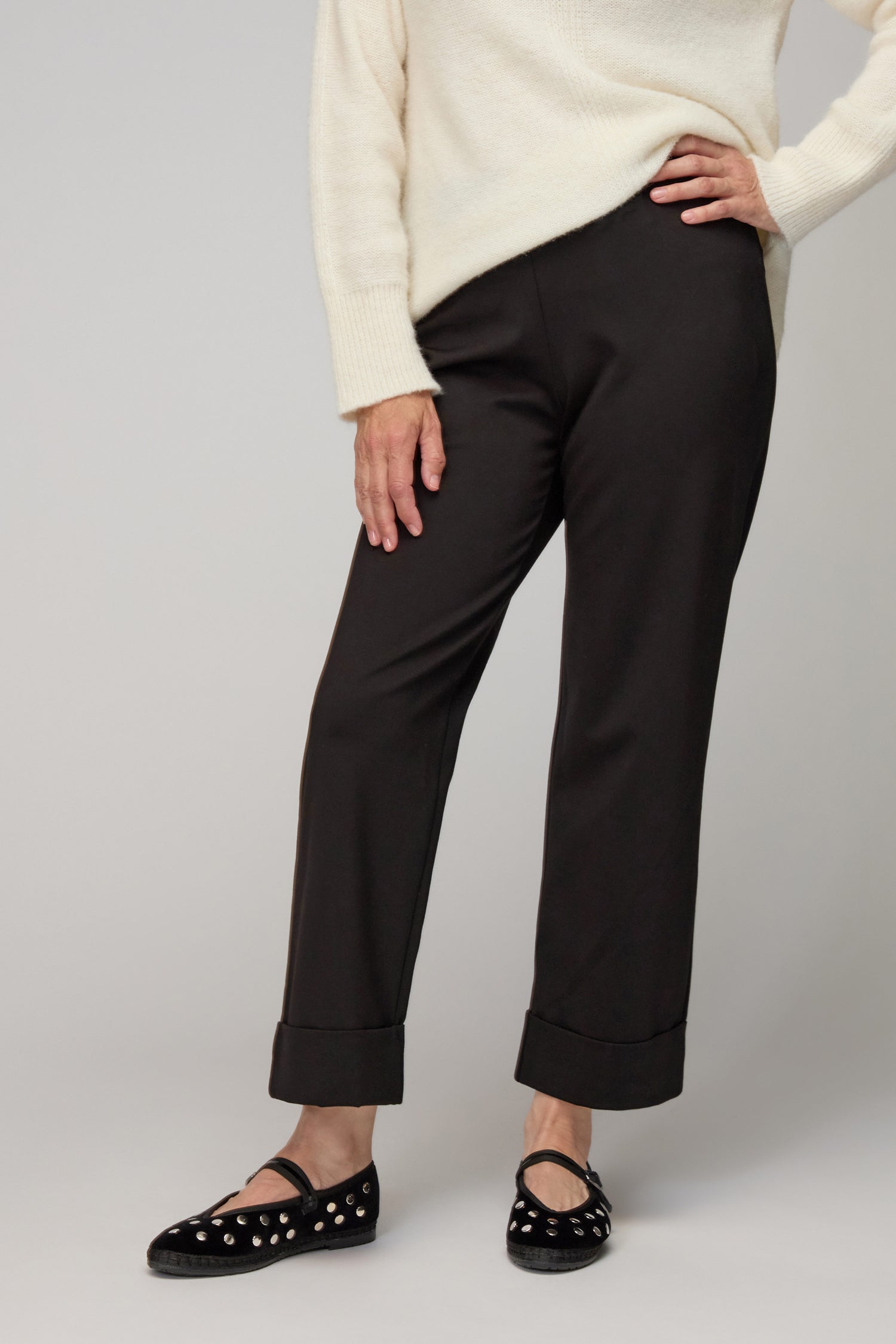 A person wearing a cream sweater, Black Ponte Straight Trousers with cuffs, and black flats with silver studs stands against a plain background.