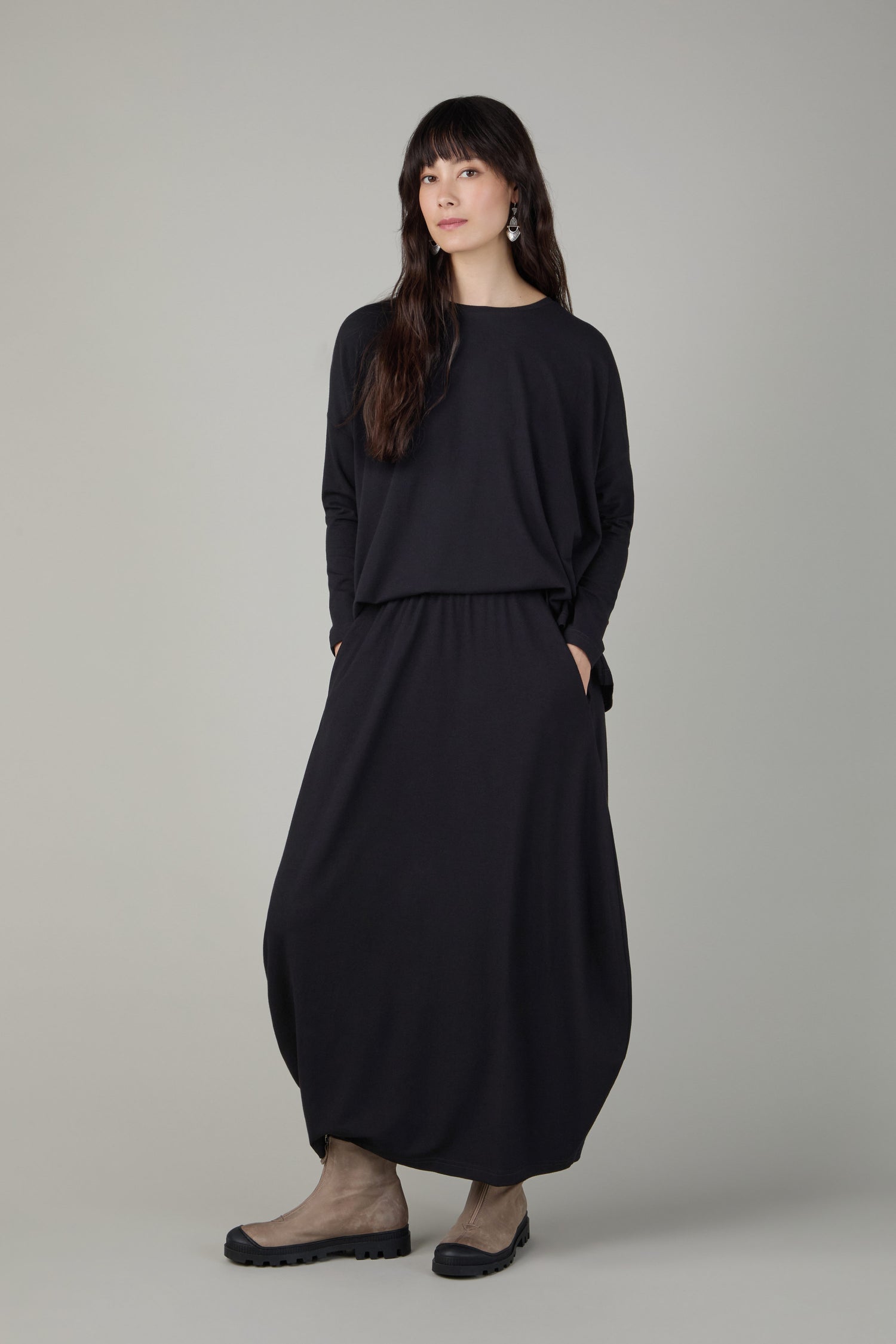 A woman stands against a plain background wearing a loose black top and the Fluid Crepe Black Bubble Skirt, styled with tan shoes and her hands in her pockets.