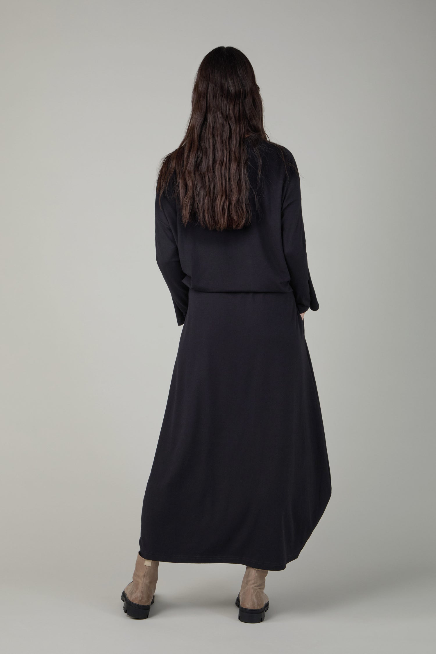 A person with long brown hair, wearing a Fluid Crepe Black Bubble Skirt and light brown boots, stands facing away from the camera against a plain background.
