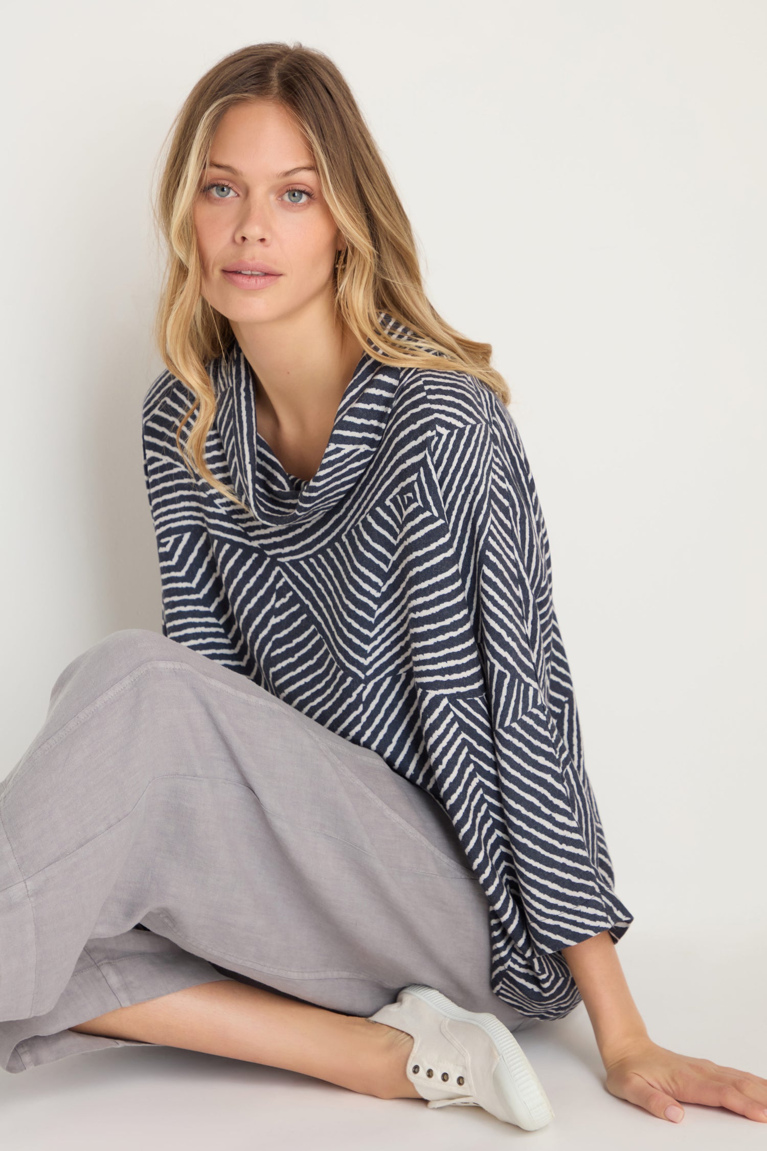 A woman with wavy blonde hair sits on the floor against a plain background, wearing the Linear Prism Lines Cowl Neck Top in navy and white brushed viscose, paired with light gray wide-leg pants and white slip-on sneakers.