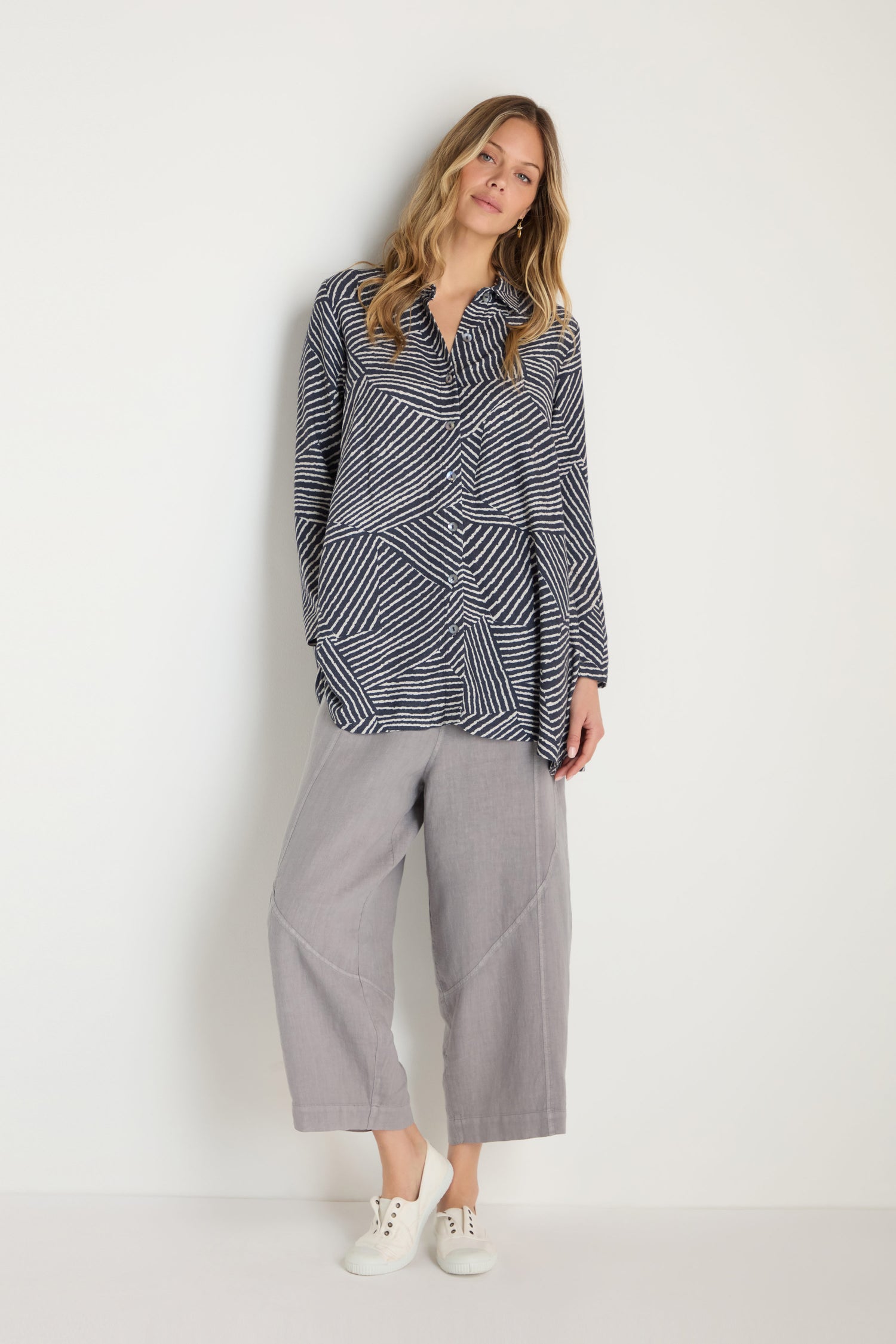 A woman stands against a plain wall wearing the Linear Prism Shirt with a loose, geometric print and A-line silhouette, paired with light gray wide-leg pants and white sneakers.