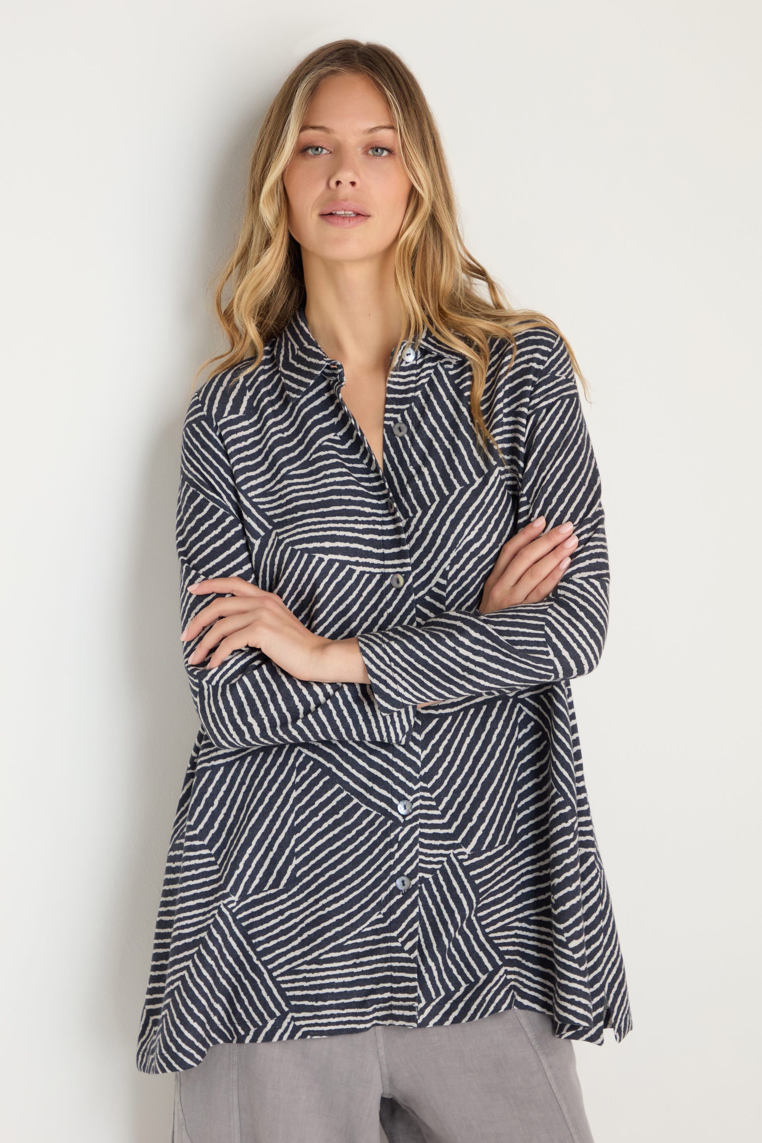 A woman stands against a light background, arms crossed, wearing the Linear Prism Shirt—a long-sleeve top with black and white geometric stripes—paired with light gray pants.