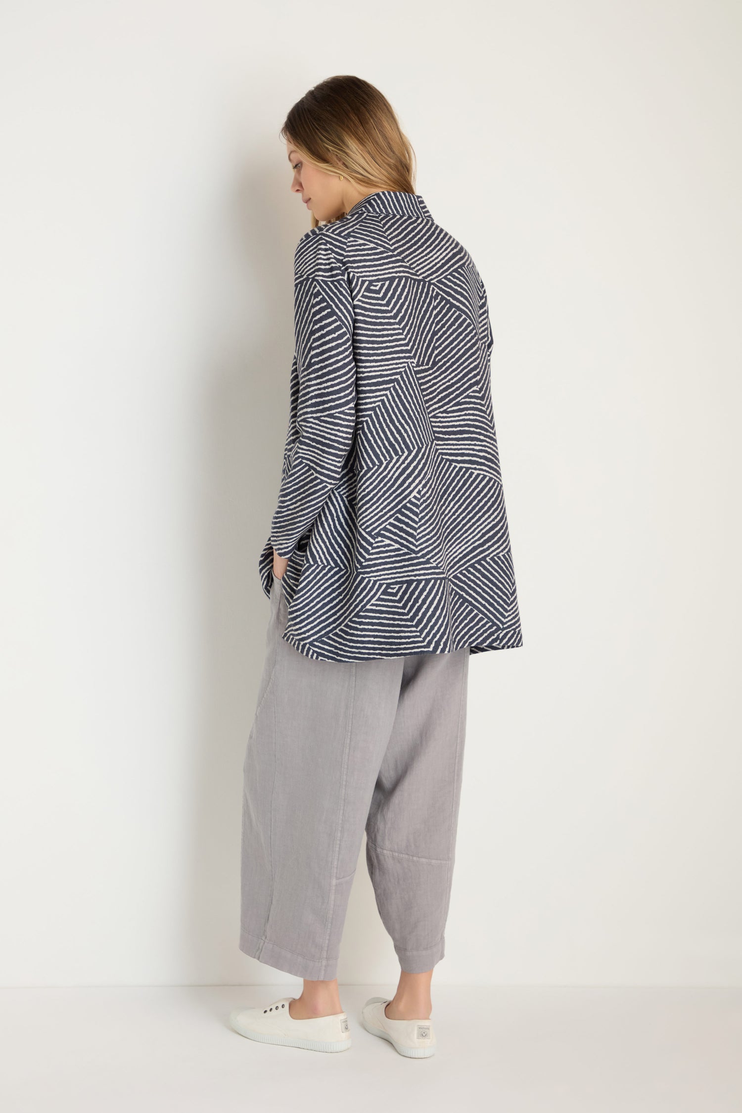 A person faces away from the camera, wearing the Linear Prism Shirt styled as a jacket, loose gray A-line pants, and white slip-on shoes against a plain light background.