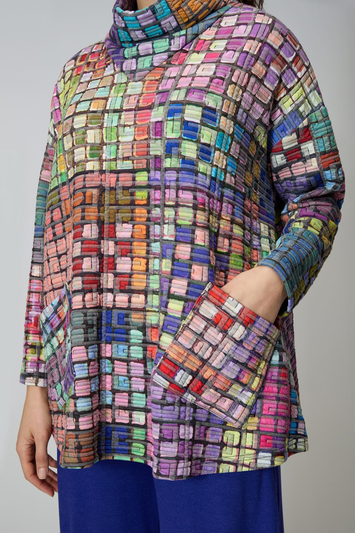 Model wears the Coloured Glass Tiles Jersey Tunic—an oversized, long-sleeve style with two front pockets and a multicolored grid pattern—paired with blue pants, shown from shoulders to mid-thigh.