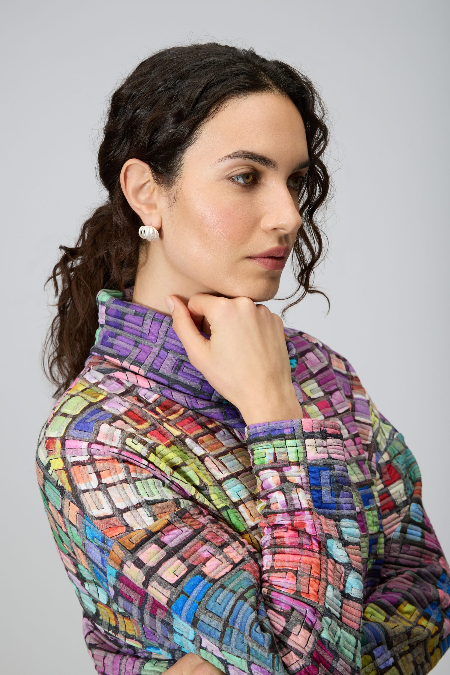 A woman with dark curly hair poses in profile, wearing the vibrant Coloured Glass Tiles Jersey Tunic with a hand near her chin against a plain background.