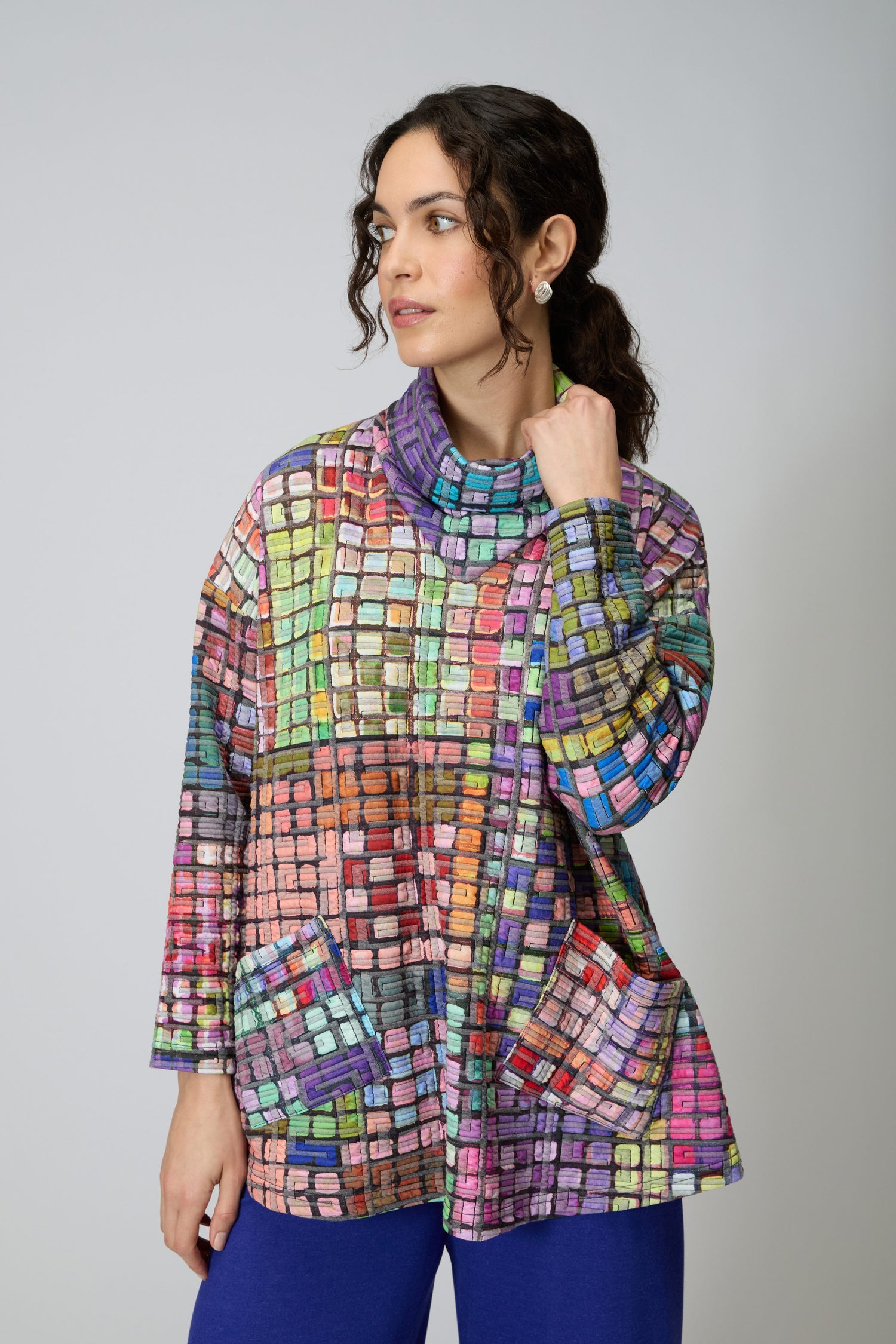 A woman wears the Coloured Glass Tiles Jersey Tunic—an oversized, grid-patterned design with large pockets—paired with blue pants against a plain light background.