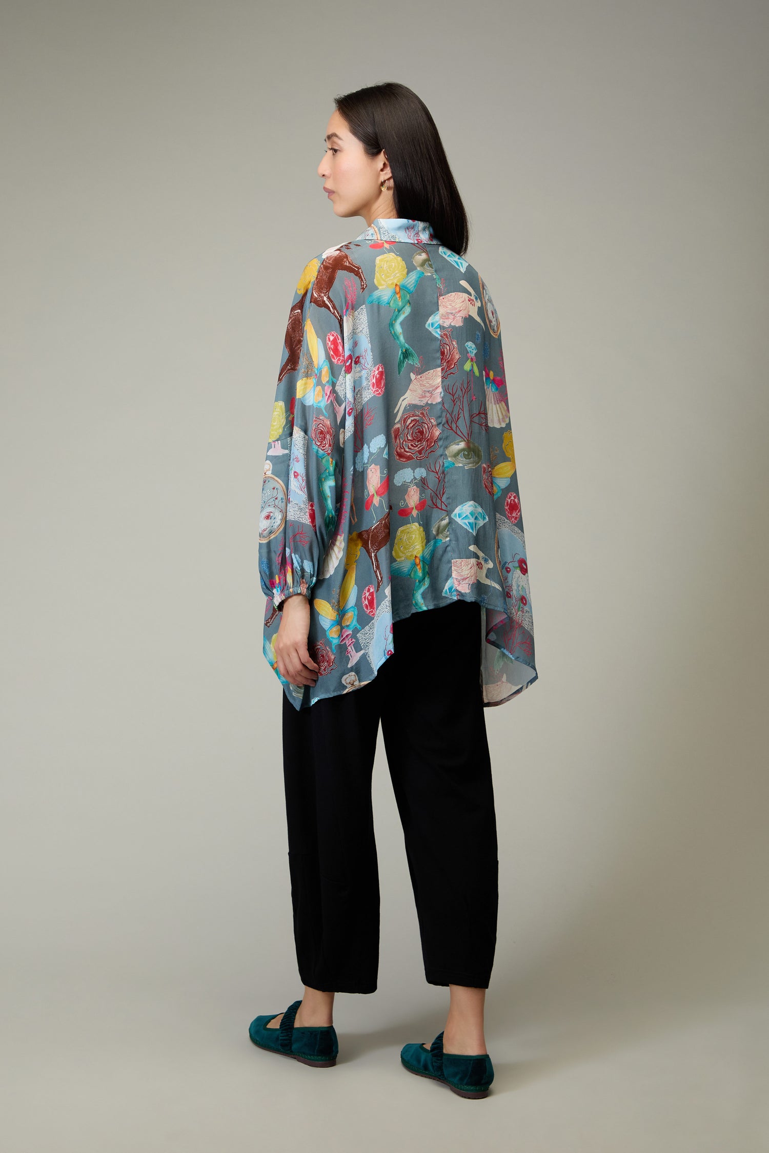 A woman faces away, wearing the Fantasy Print Shirt with a loose, colorful floral pattern in a Yavi illustrative style, paired with black pants and teal shoes against a plain background.
