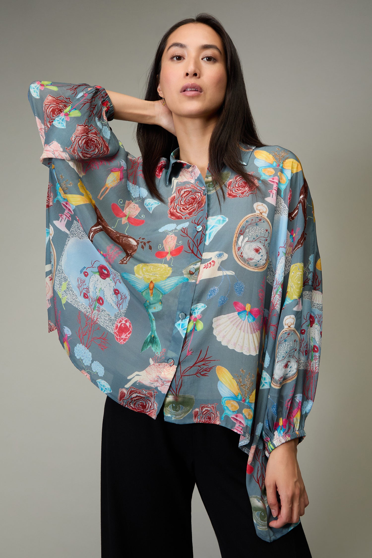 A woman in black pants wears the Fantasy Print Shirt, featuring a whimsical collage of flowers, birds, and objects in Yavi's illustrative style. She stands against a plain gray background.