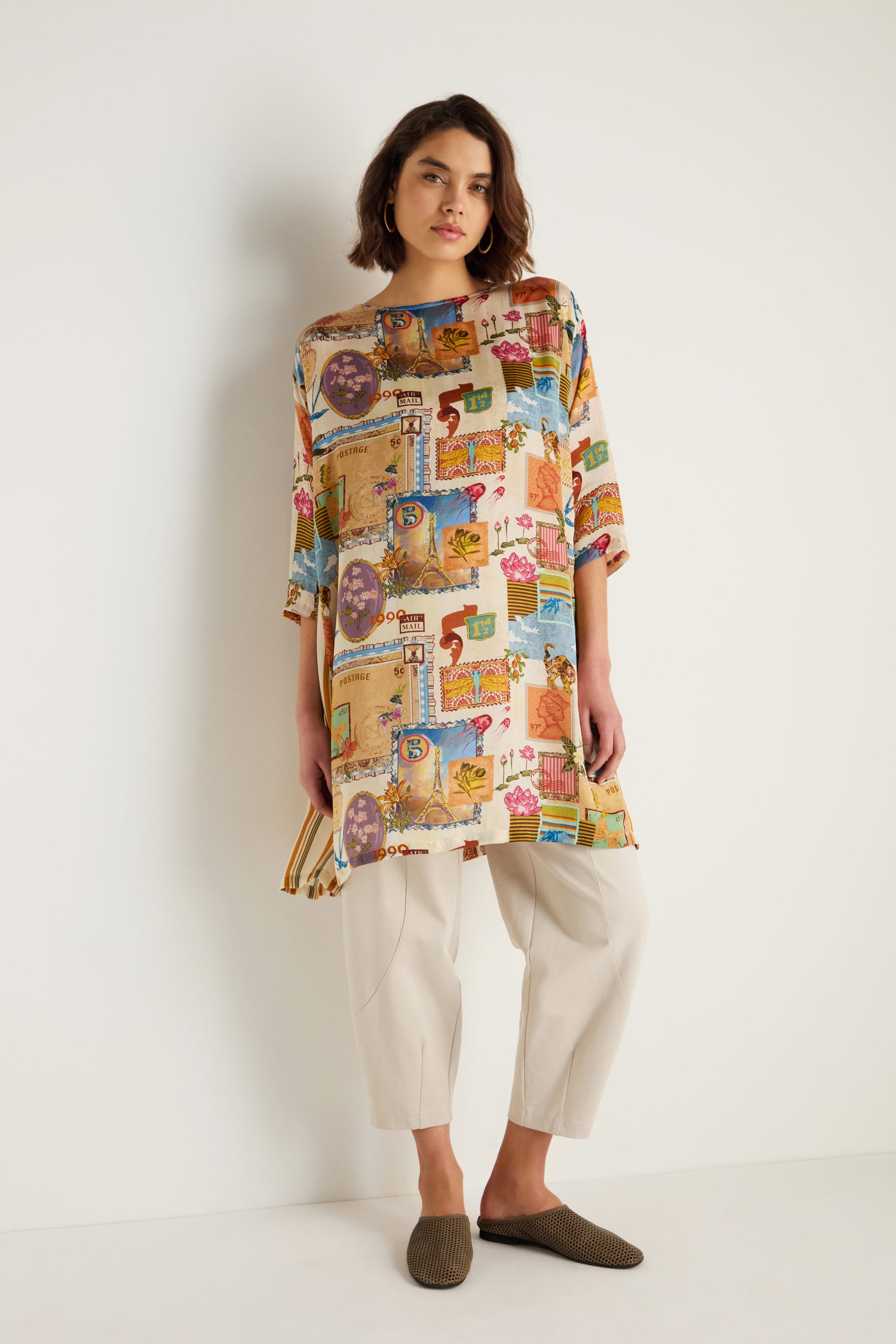 A woman stands against a white wall wearing the Postcard Print Cupro Tunic with vibrant hand-drawn prints, paired with light beige pants and brown slip-on shoes.