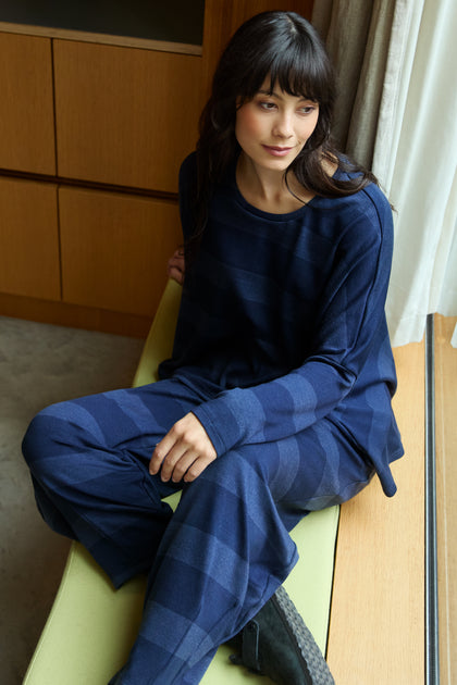 A person with long dark hair sits on a cushioned bench by a window, wearing the Soft Viscose Jersey Stripe Boxy Top and black shoes, the loose fit draping comfortably for a relaxed look.