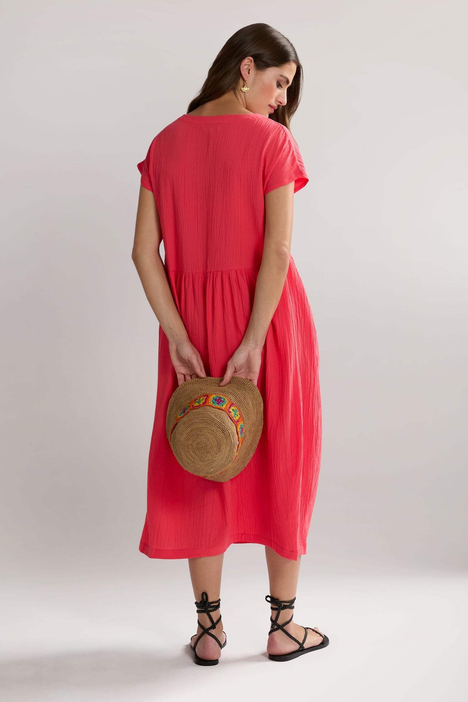 A Woman in a Crinkle Viscose Bubble Dress stands with her back to the camera, holding a straw hat decorated with a colorful band. The textured pink fabric of her midi-length skirt complements her black sandals. The background is plain white.