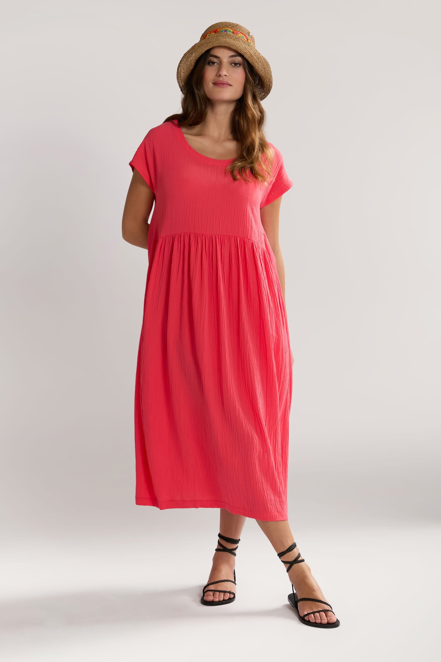 A woman stands wearing a bright pink Crinkle Viscose Bubble Dress with short sleeves, paired with black strappy sandals and a straw hat. She poses against a plain white background.