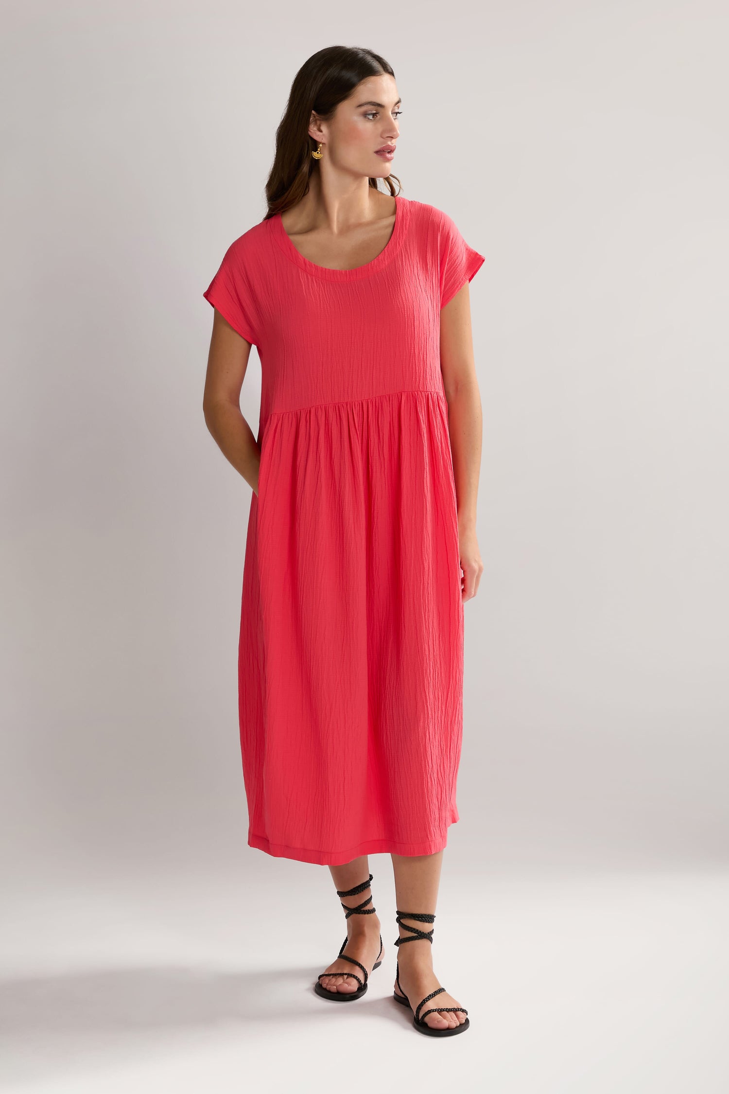 A woman stands against a plain background wearing a Crinkle Viscose Bubble Dress in bright pink, featuring short sleeves and a midi-length skirt, paired with black strappy sandals.