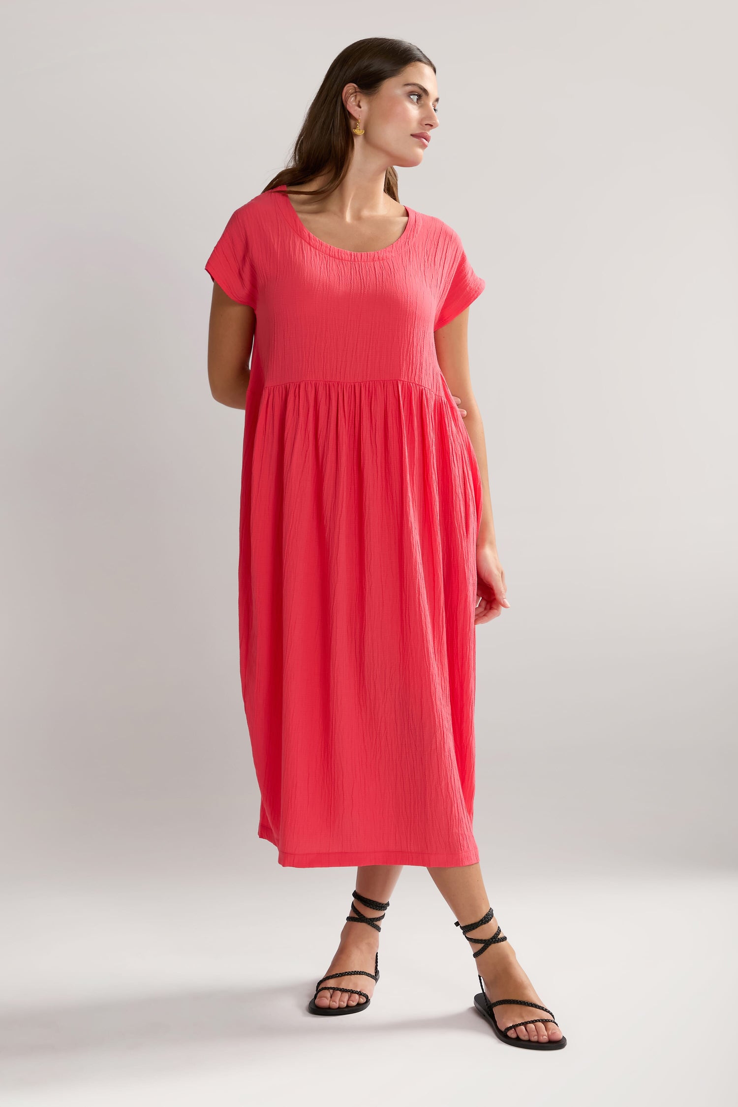 A woman stands wearing a loose-fitting, Crinkle Viscose Bubble Dress with a midi-length skirt and strappy black sandals. She looks slightly to the side against a plain gray background, the textured pink fabric adding subtle depth to her coral-colored outfit.