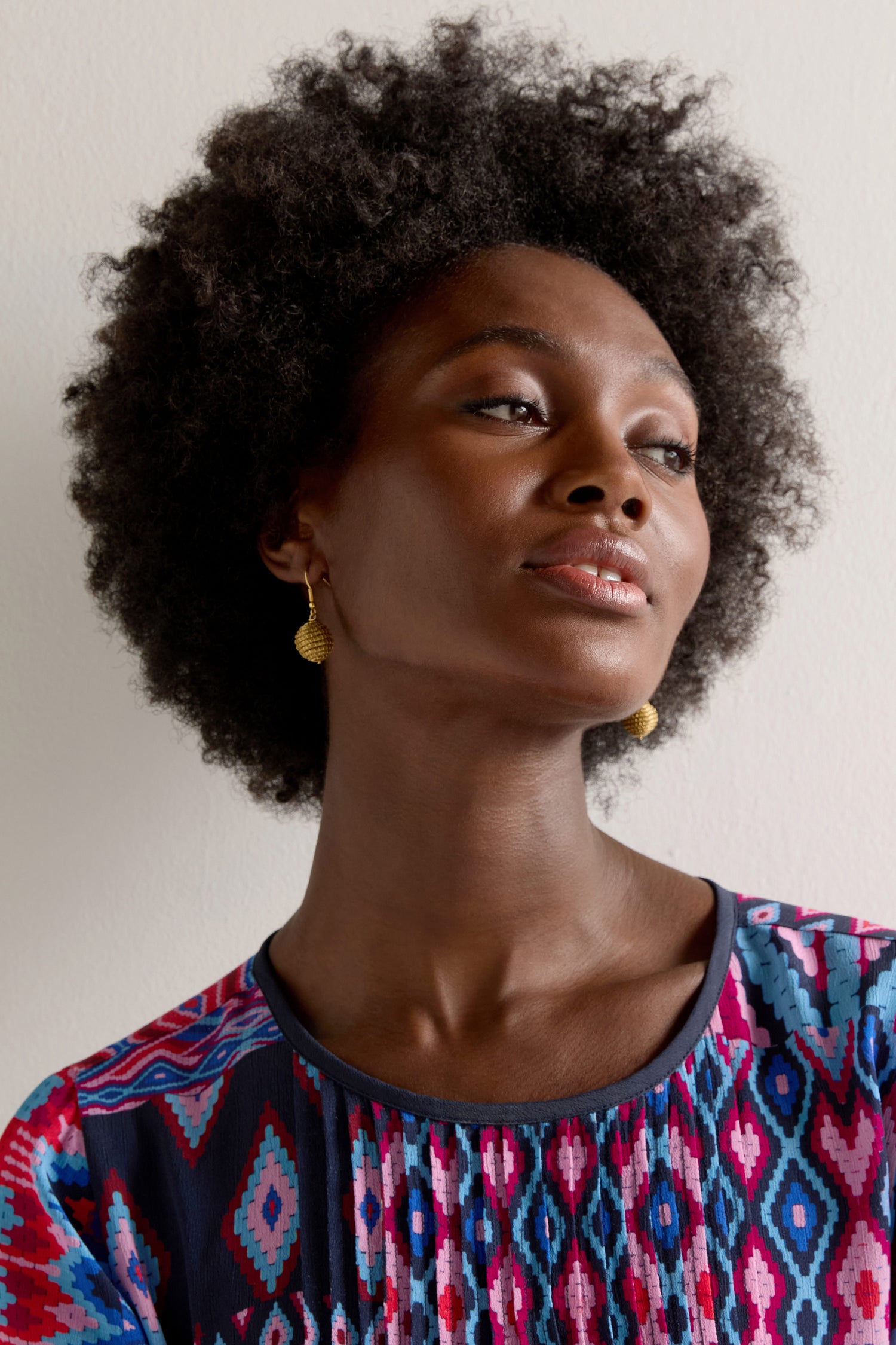 Wearing Golden Grass Woven Sphere Earrings and a vibrant patterned top, a person with naturally curly hair looks upward against a plain background.