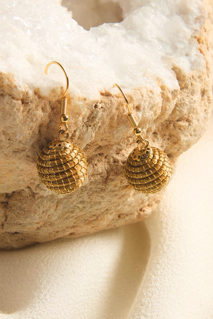 The Golden Grass Woven Sphere Earrings feature a textured design, gracefully suspended from hooks on a textured stone-like surface, offering a handcrafted artisan jewelry look.