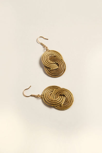 The Golden Grass Woven Circles Earrings feature two gold-tone dangles with interlocking spiral designs on a light background, embodying the essence of handcrafted sustainable accessories.