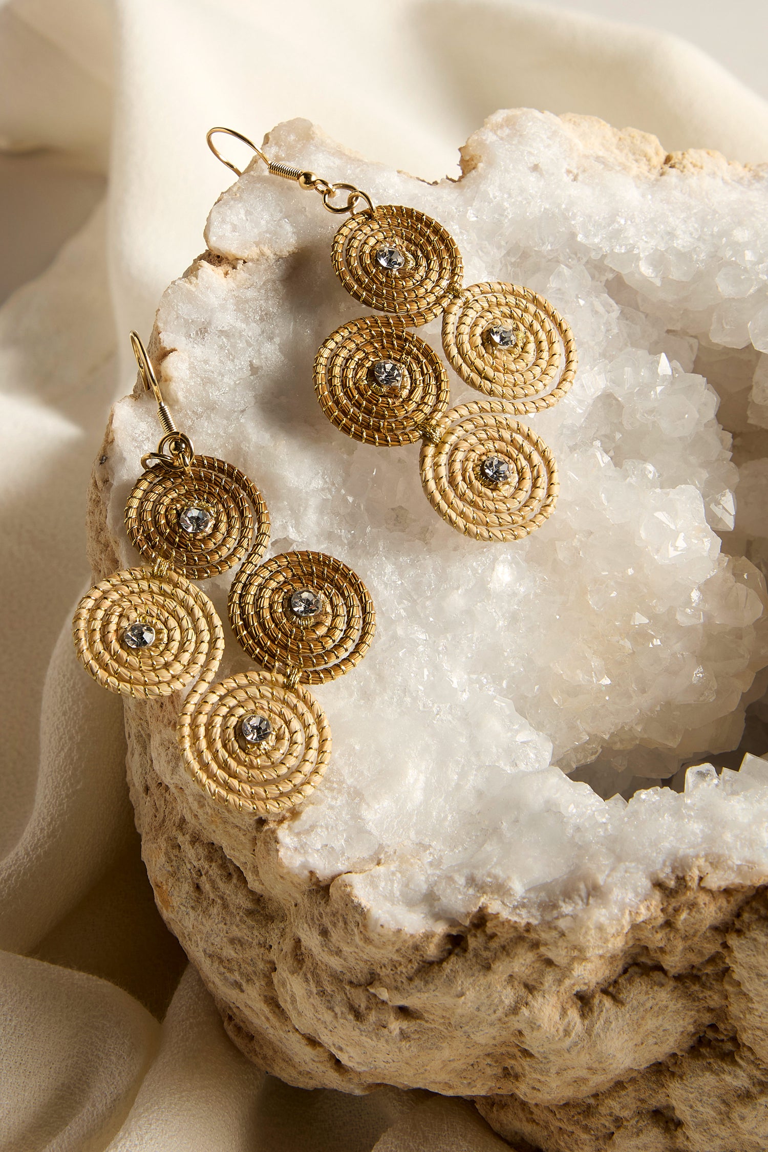 The Golden Grass Woven Spiral Rhinestone Earrings are elegantly showcased on a crystal geode and light fabric, capturing the essence of sustainable jewelry.