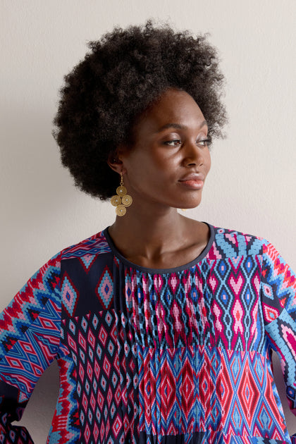 A woman with an afro hairstyle, wearing a colorful geometric-patterned shirt and Golden Grass Woven Spiral Rhinestone Earrings, looks to the side against a plain background.