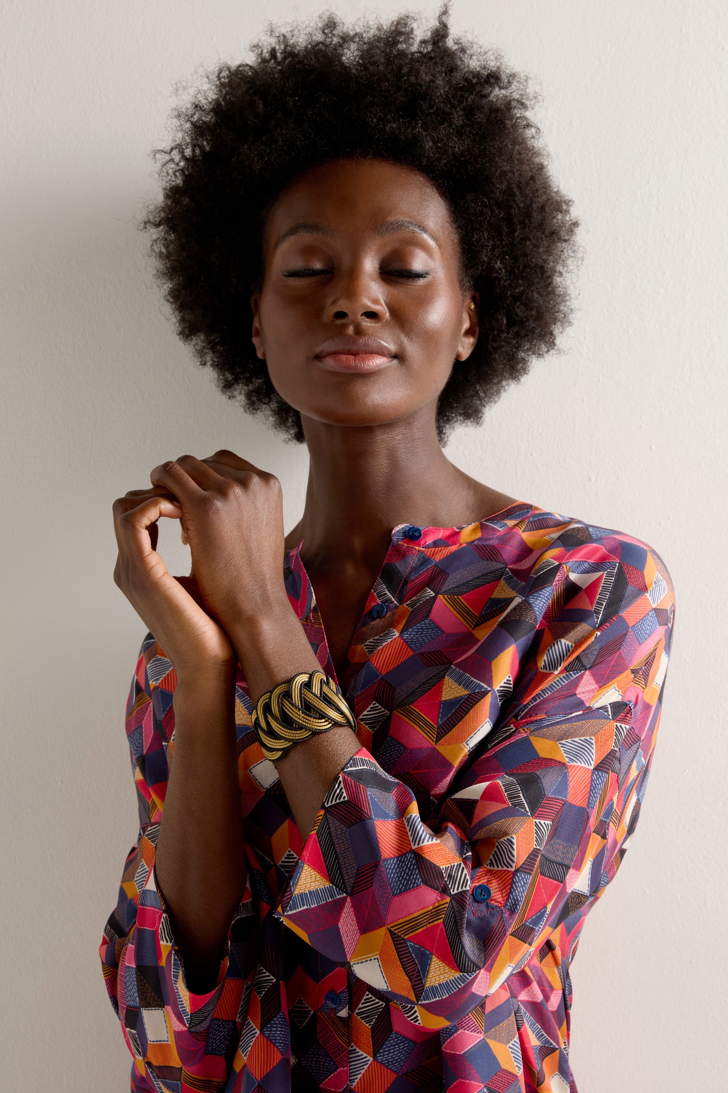 With eyes gently closed, the person stands against a plain backdrop wearing a colorful geometric patterned top and a Gold and Black Grass Woven Bracelet.